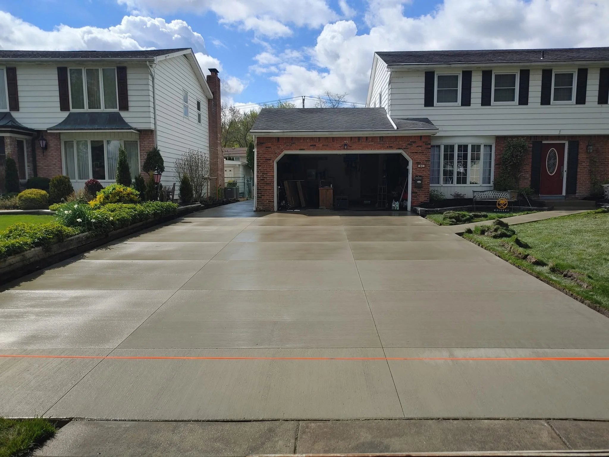 A residential driveway leading to a brick garage, with a two-story house on each side and well-maintained gardens, under a partly cloudy sky.