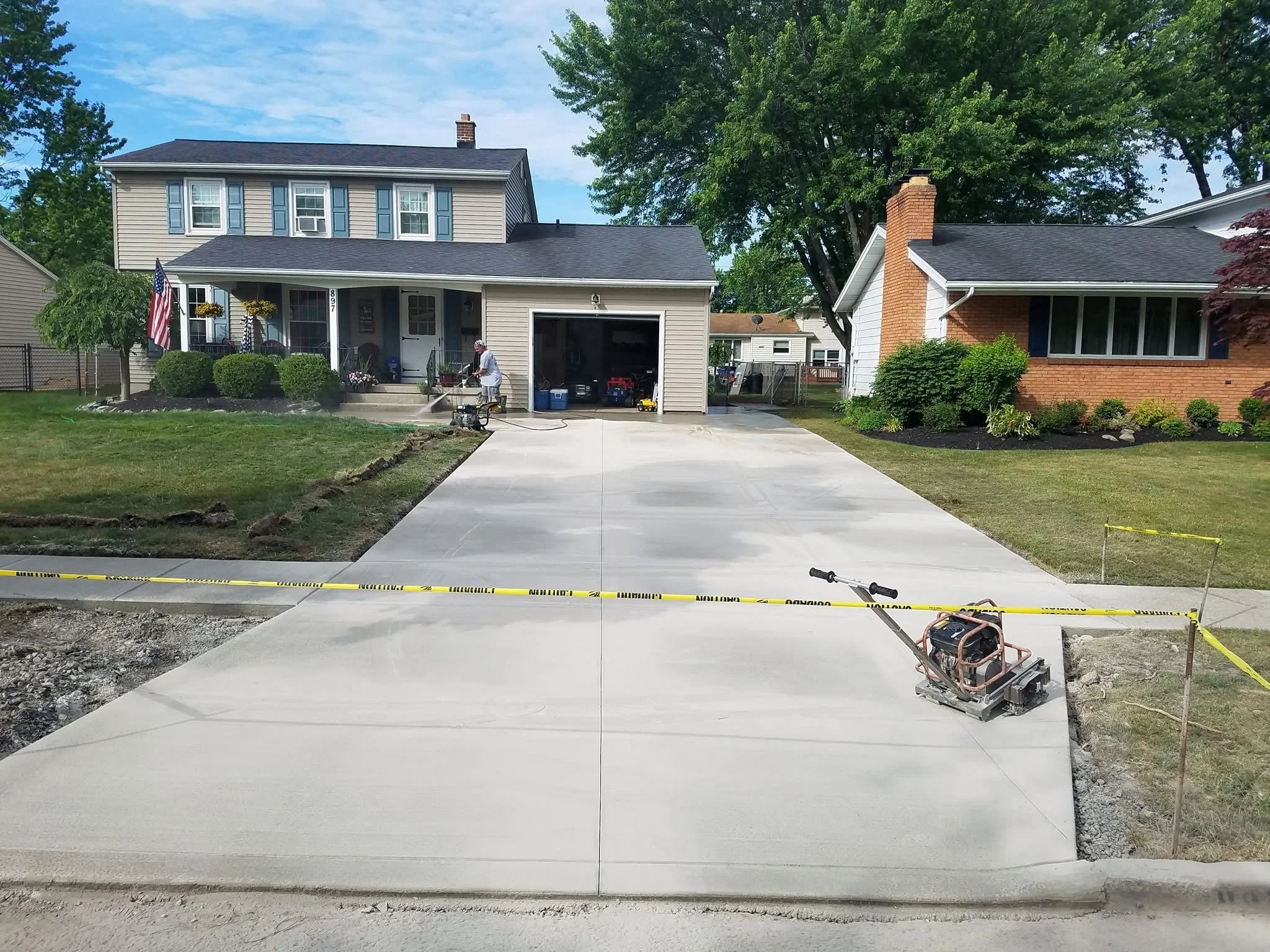 Newly poured concrete driveway in front of two houses, with a worker smoothing the concrete with a power screed, and construction tape marking the area, during daytime.