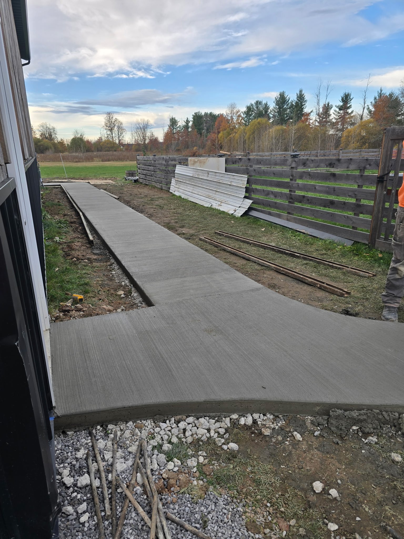 Newly poured concrete sidewalk being constructed outside a building, with construction materials and a partially finished fence in the background, under a partly cloudy sky.