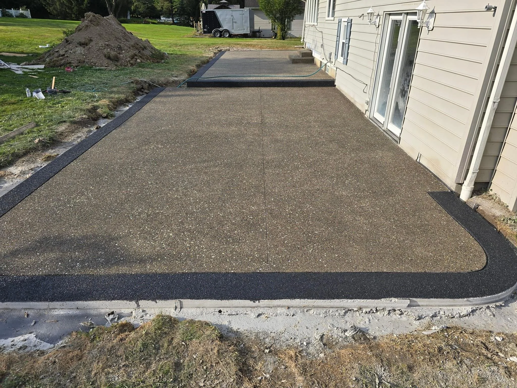 Freshly poured concrete patio with black border in backyard, adjacent to house with sliding glass door, with yard and trees in background.