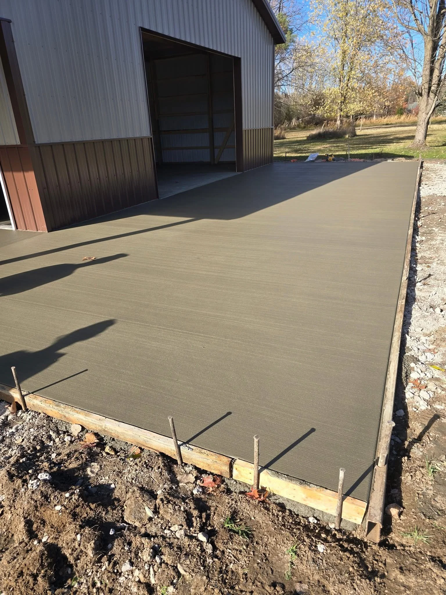 Freshly poured concrete slab outside a metal building, with wooden formwork and metal stakes, in a yard with trees and grass.