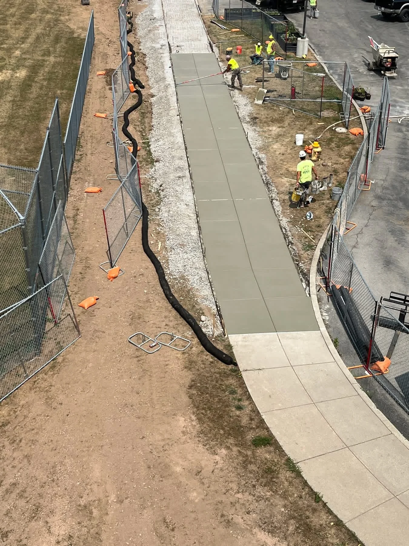 Construction workers are building a sidewalk, with some pouring concrete and others using tools. Fencing surrounds the work site.