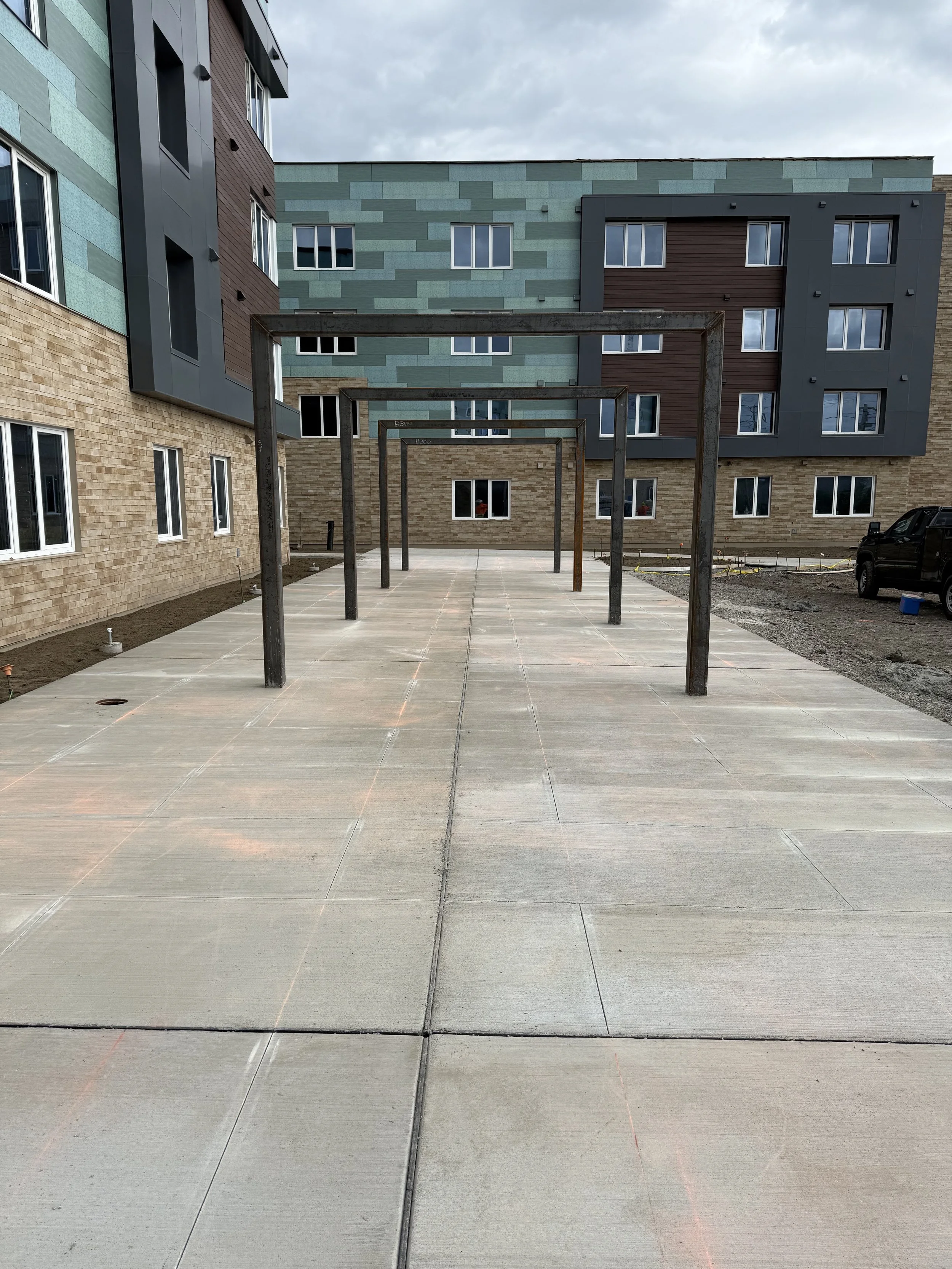 Concrete sidewalk with wooden posts and trim marks in front of a multi-story apartment building with various colored panels.