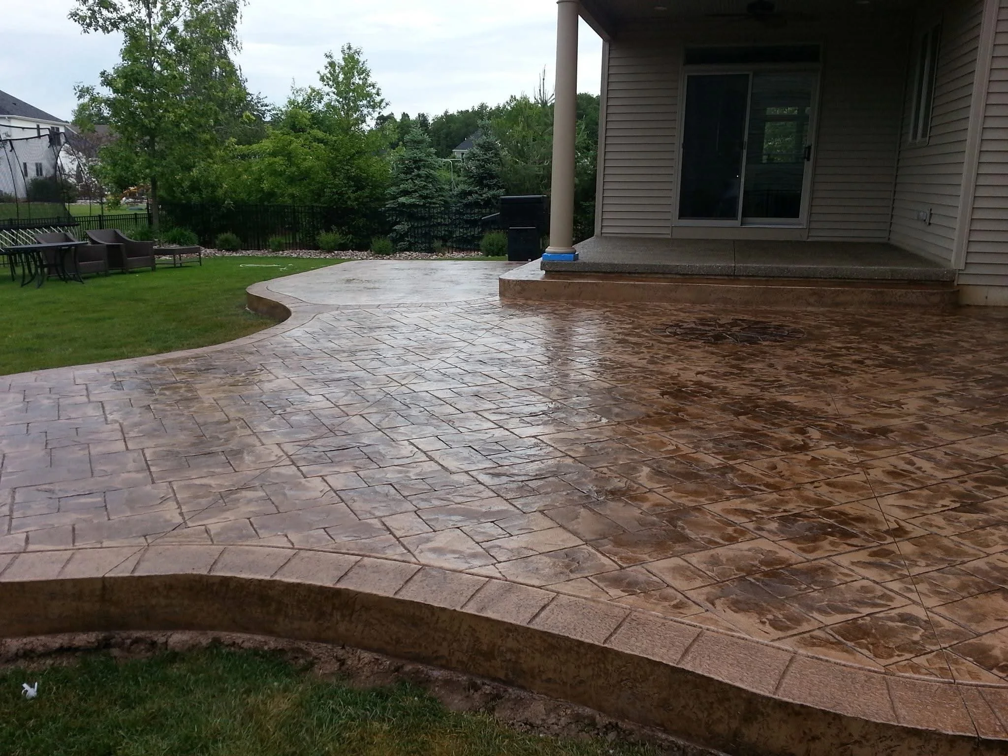 Wet stamped concrete patio extending from the house to a grassy yard with outdoor furniture, trees, and a black fence in the background.