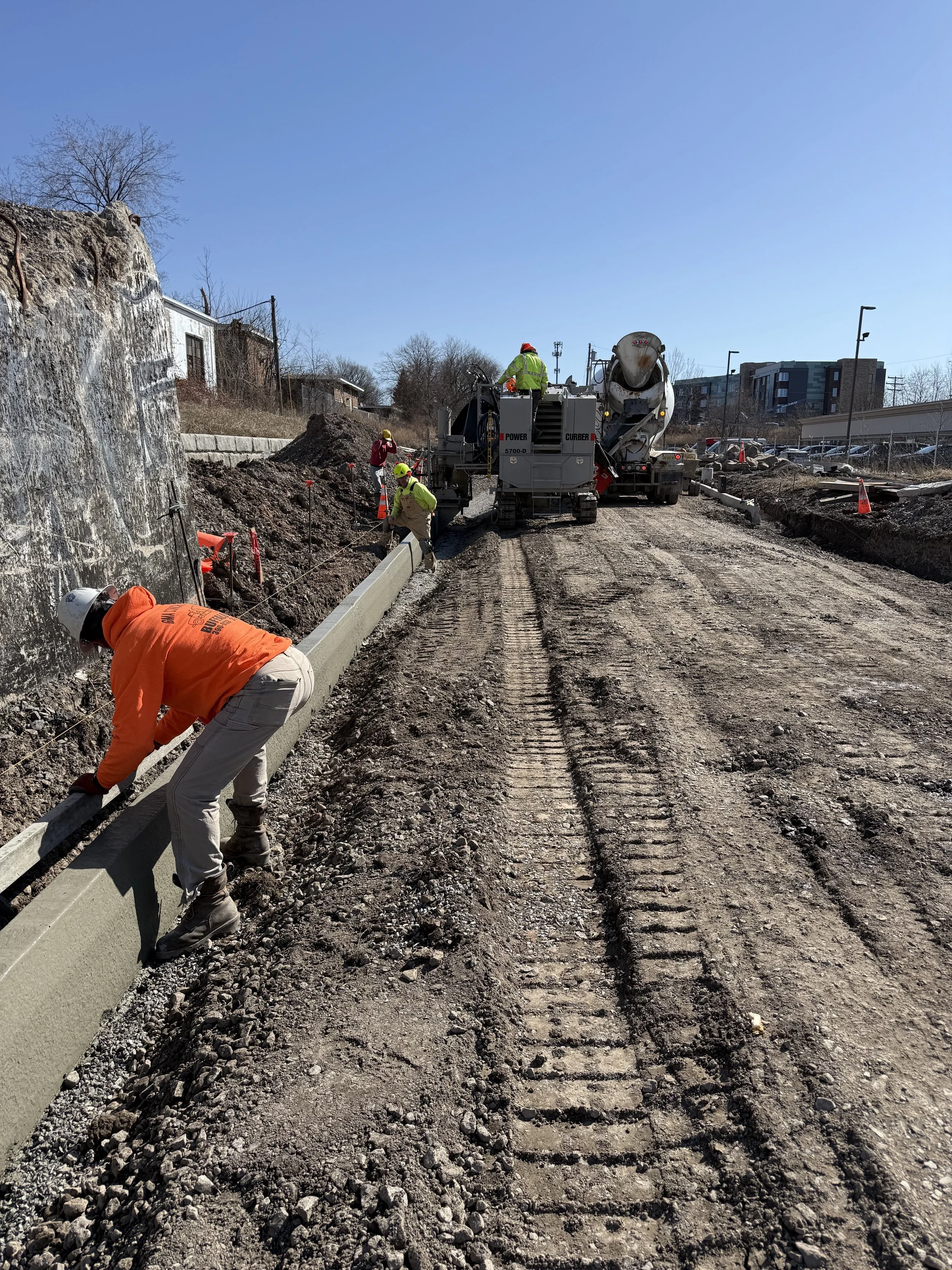 Construction workers installing a curb alongside a dirt road with construction machinery in the background.