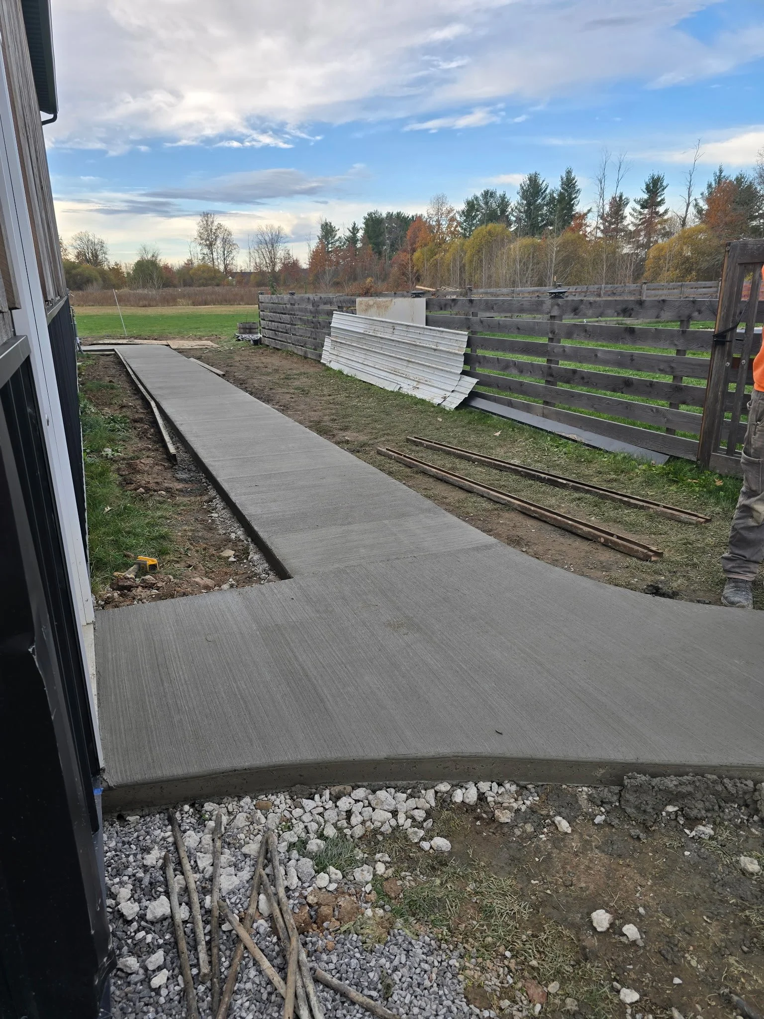 Newly poured concrete sidewalk with a gentle curve in a backyard, surrounded by gravel, dirt, and construction materials, with a wooden fence and trees in the background under a partly cloudy sky.