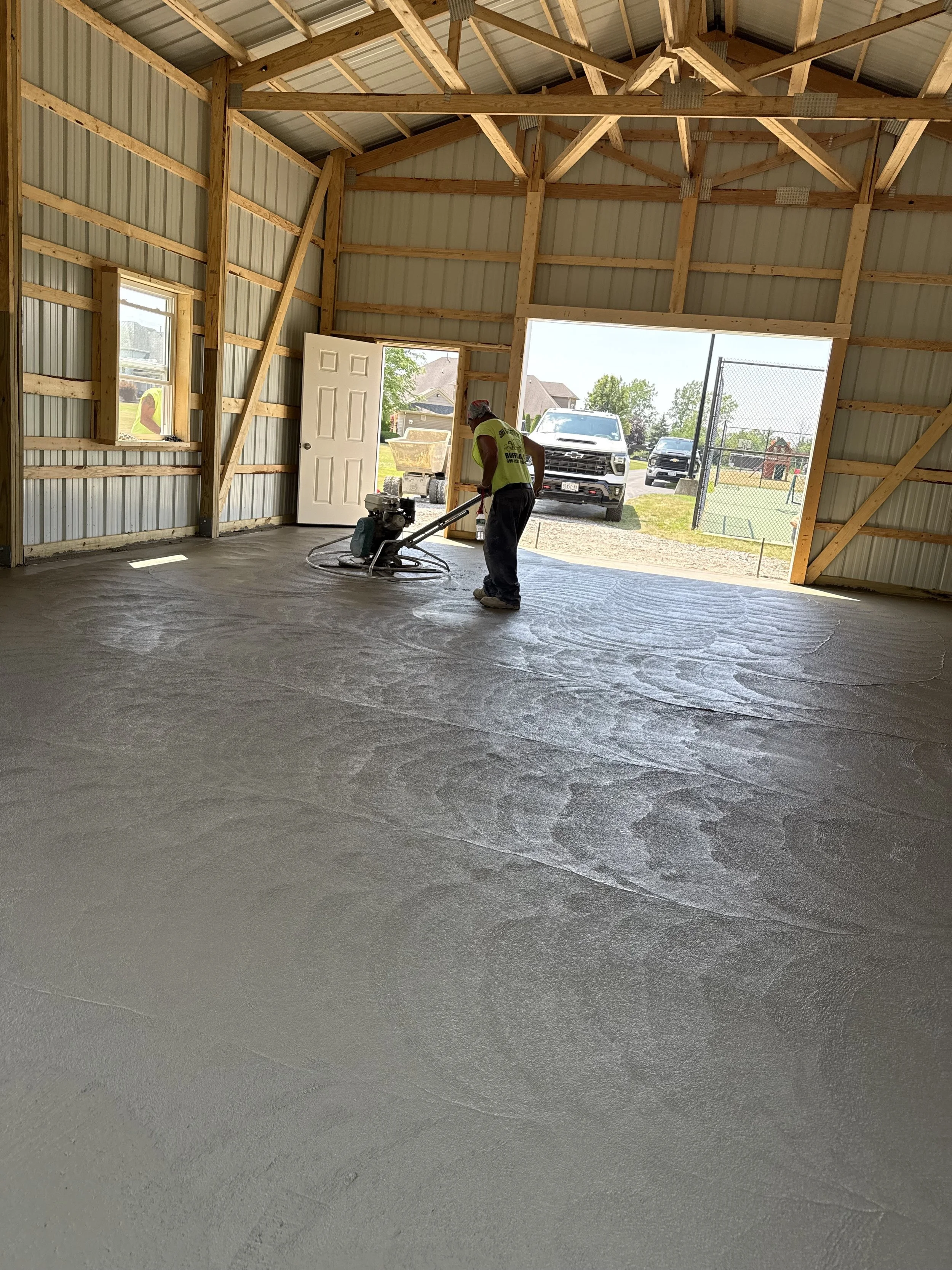 Worker smoothing freshly poured concrete floor inside a wooden barn with open garage door, revealing cars outside and a tennis court.