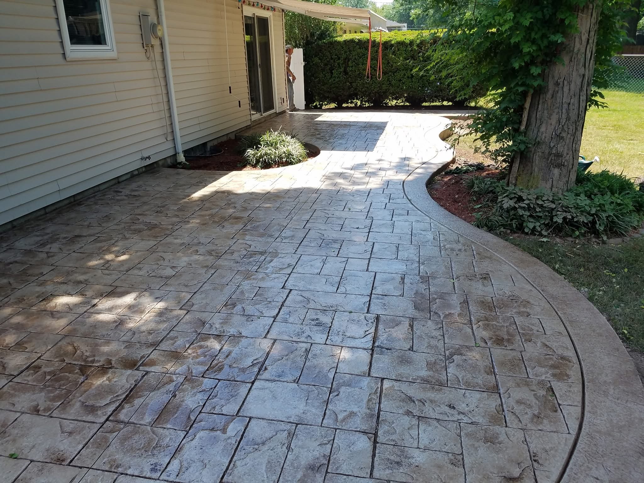 Newly paved backyard patio with stamped concrete and curved border, next to a house with beige siding, a tree, and grass lawn.