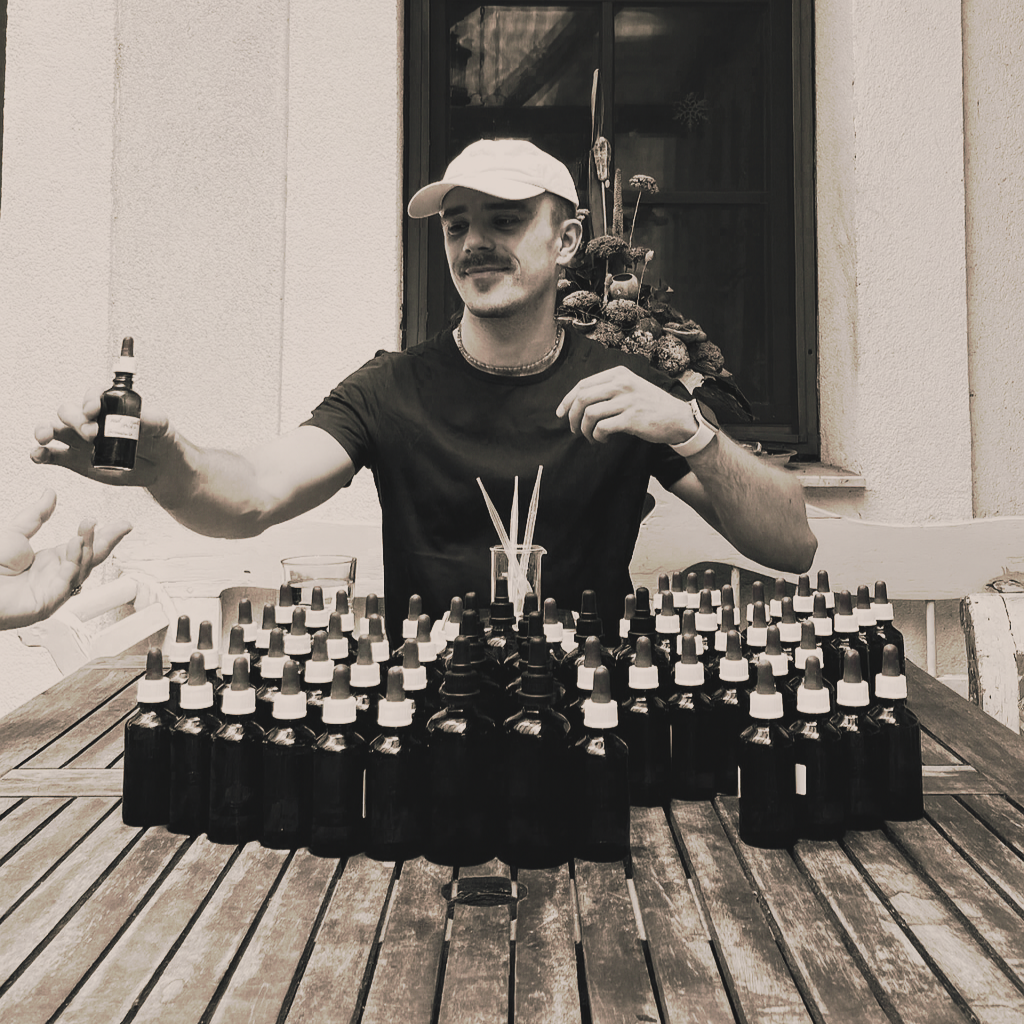 A man in a white cap and black shirt handing a small bottle to someone across a wooden table, which has multiple bottles with dropper caps and some sticks in a jar, outside in front of a window with decorative flowers in the background.