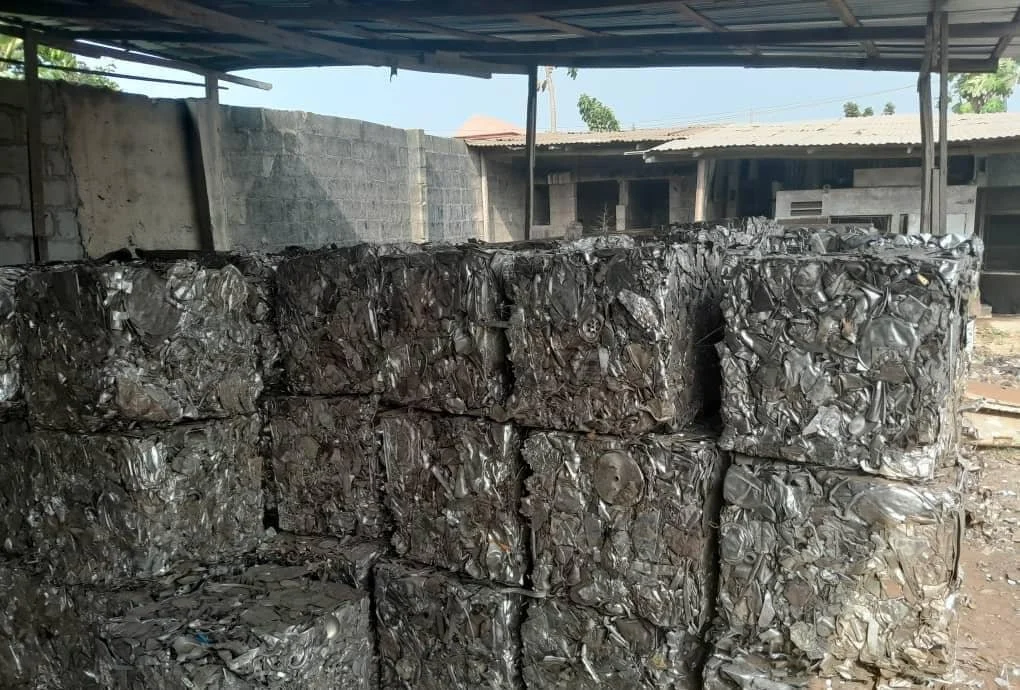Stacks of compressed scrap metal outside under a roof in a yard surrounded by concrete and wooden structures.