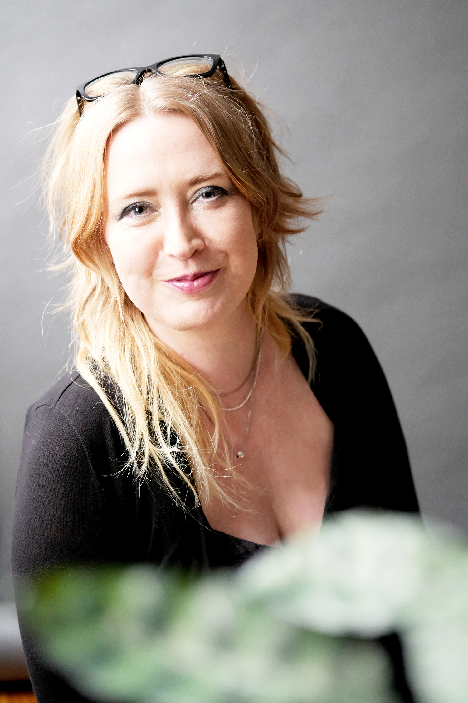 A woman with shoulder-length blonde hair, wearing glasses on her head, a black top, and jewelry, smiling at the camera against a gray background.