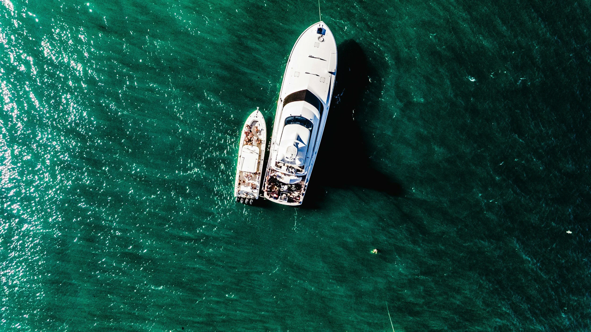 An aerial view of two large white yachts floating on turquoise water, casting a shadow in the water.