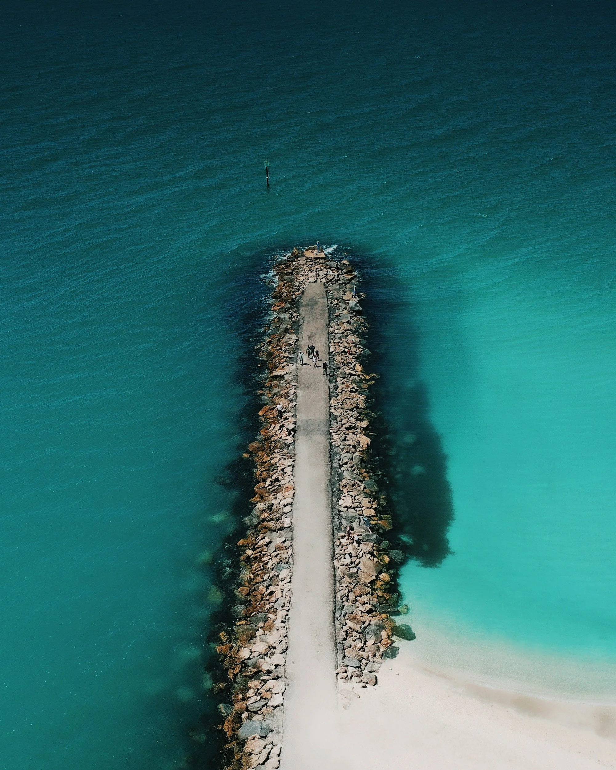 An aerial view of a narrow stone pier extending into the ocean, with two people walking on it, and the shoreline with turquoise water and white sand at the end.