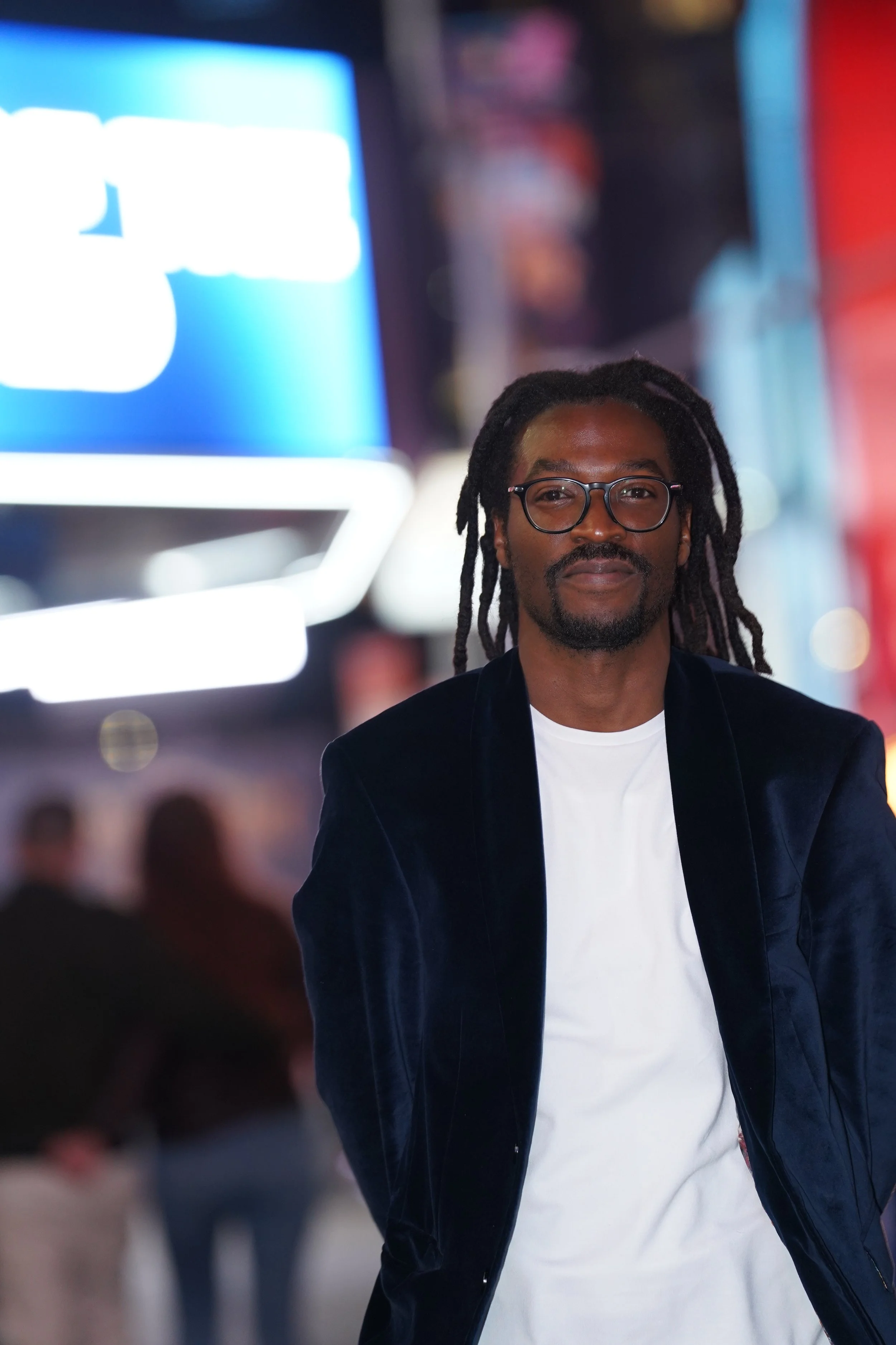 A man with glasses and dreadlocks standing in a city at night, illuminated by bright neon lights and billboards.
