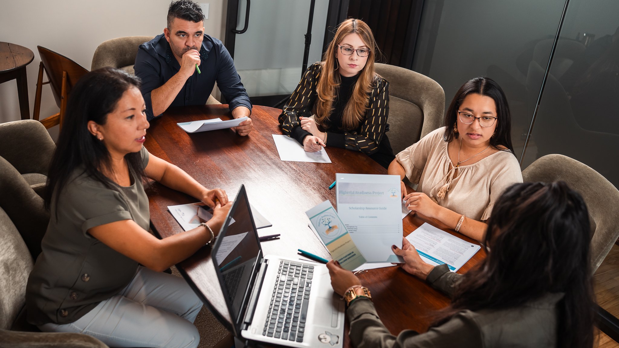 A group of six diverse women and one man sitting around a wooden conference table in a business meeting, discussing documents and using a laptop.