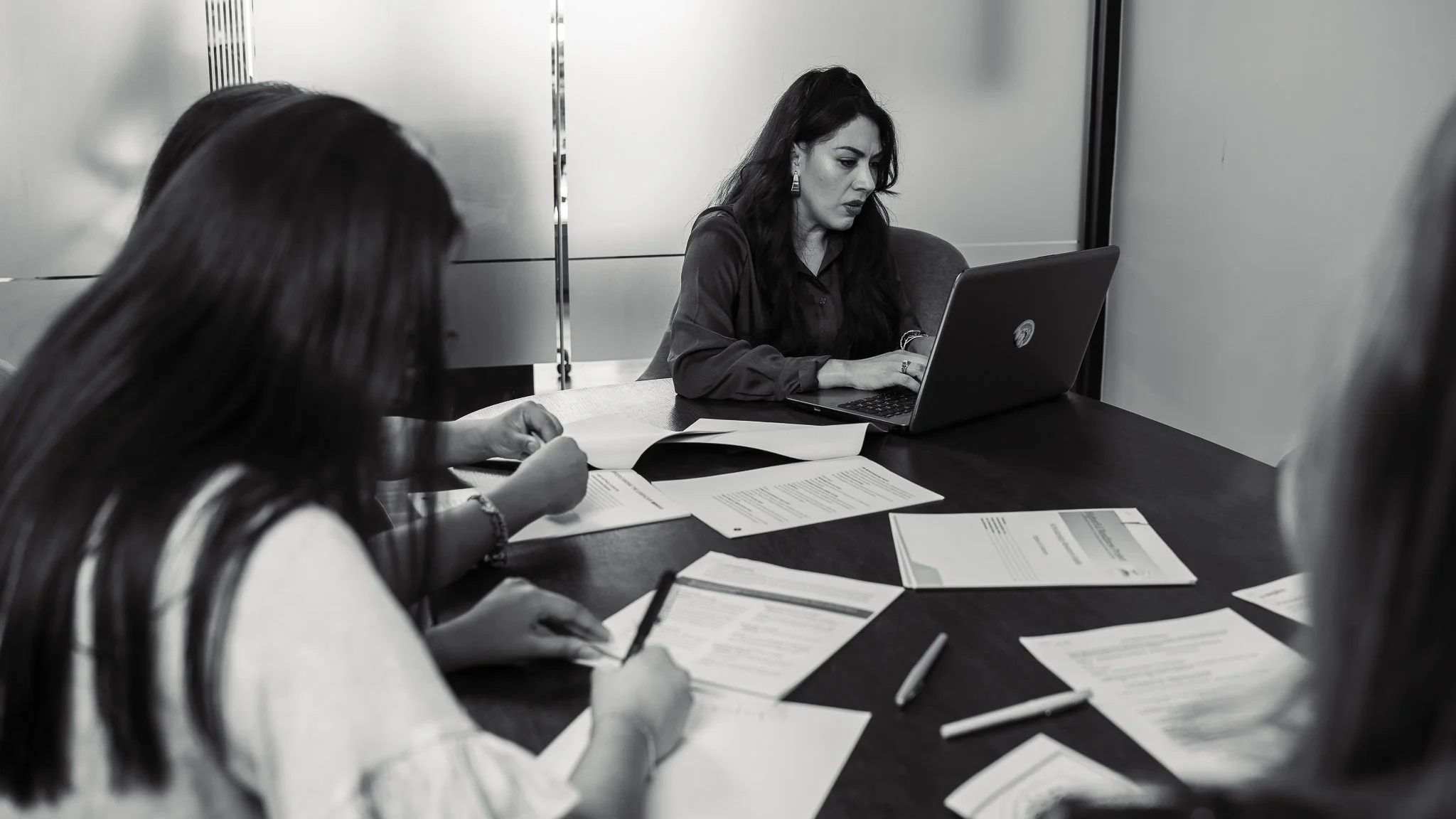 Three women are working on documents and using a laptop in a meeting room.
