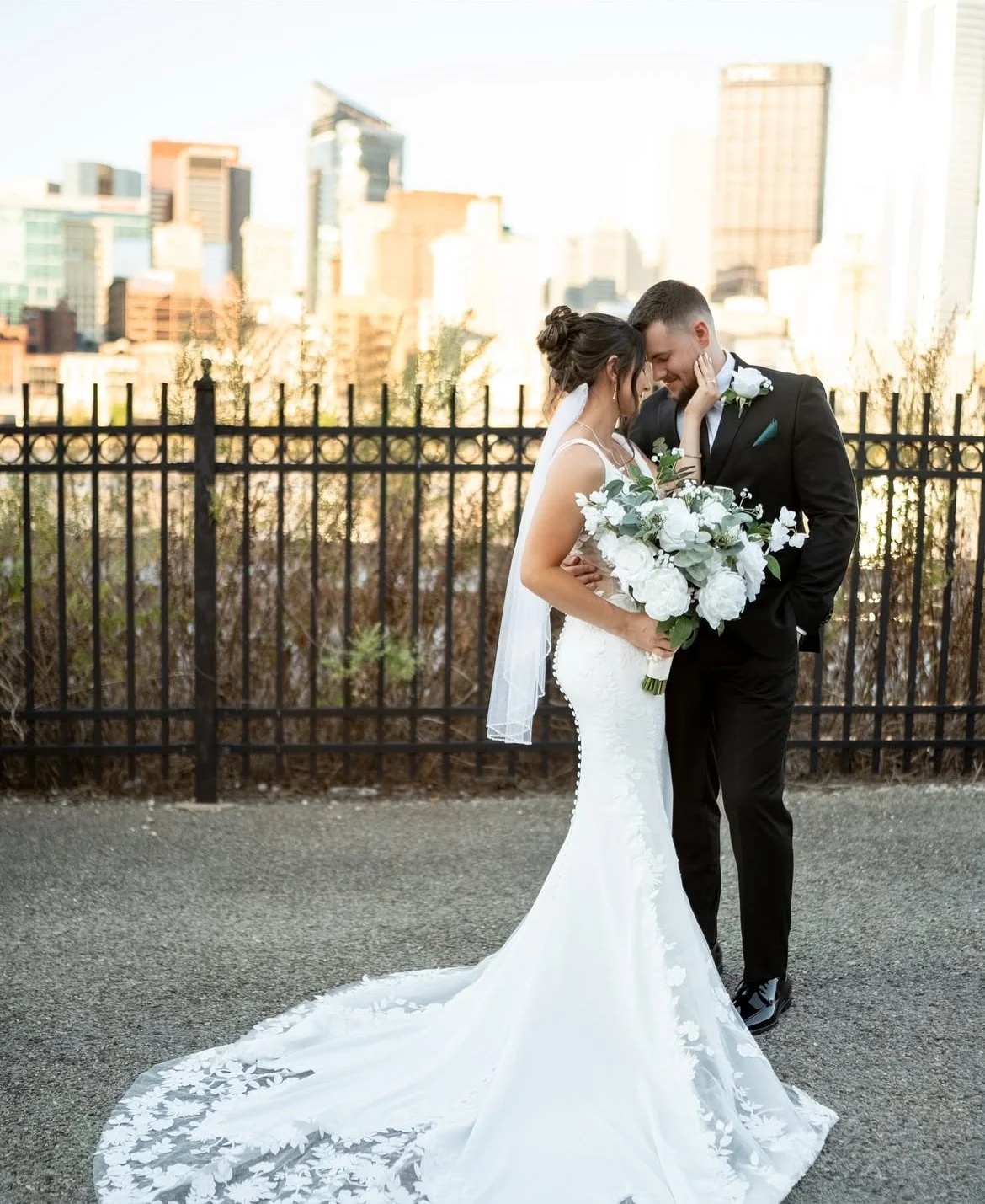 A bride and groom standing close together on a city rooftop, sharing a tender moment, with a city skyline in the background.