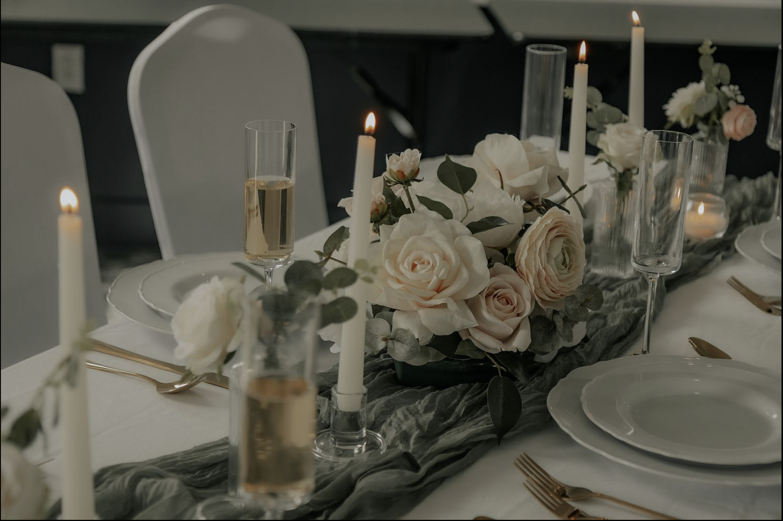 Elegant table setting with white plates, gold flatware, tall candles, champagne glasses, and a floral centerpiece of white and blush roses with greenery, on a dark and light table runner.