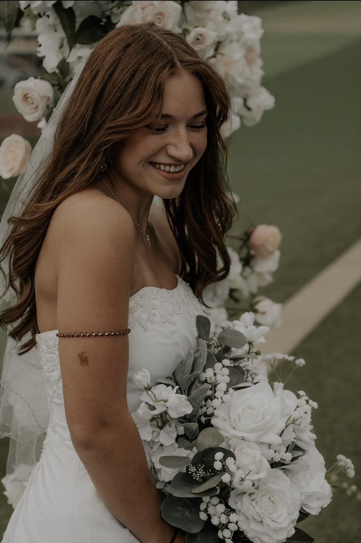 Smiling bride with long brown hair in a white wedding dress holding a bouquet of white flowers, standing outdoors near a floral display.