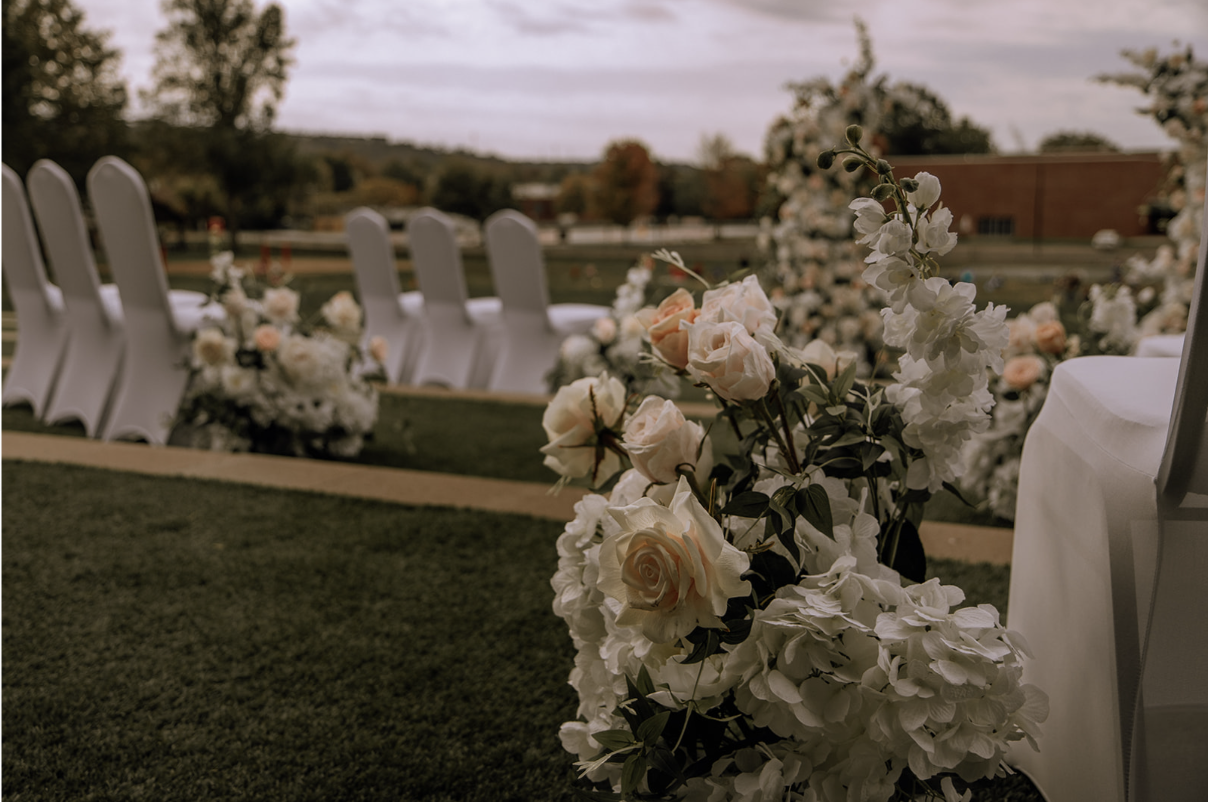 Outdoor wedding ceremony setup with white chairs arranged on grass and decorated with floral arrangements, including white roses and hydrangeas, against a backdrop of trees and a cloudy sky.