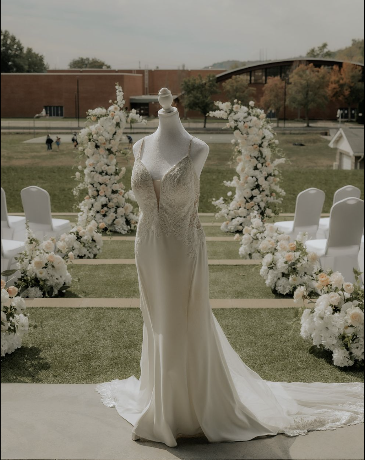 A wedding dress displayed on a mannequin in an outdoor wedding ceremony setting, surrounded by floral arrangements and chairs.
