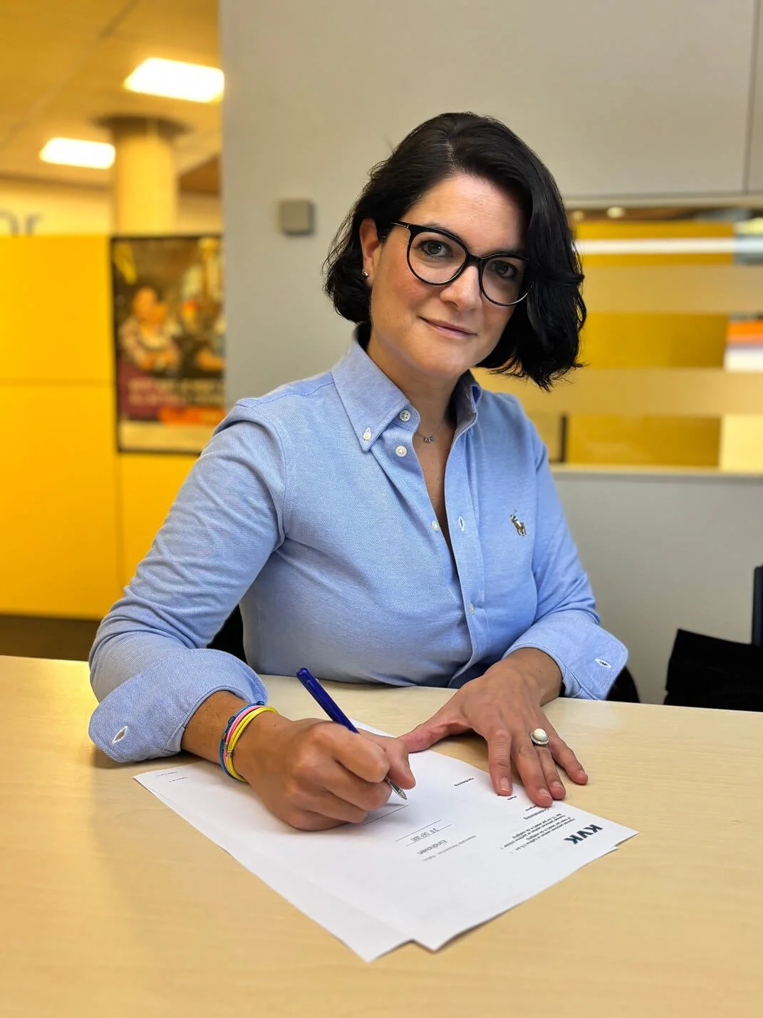 A woman with short black hair and glasses sitting at a table, signing a document with a blue pen. She is wearing a light blue button-down shirt and colorful bracelets on her wrist, in an office or reception area with yellow walls and a poster in the background.