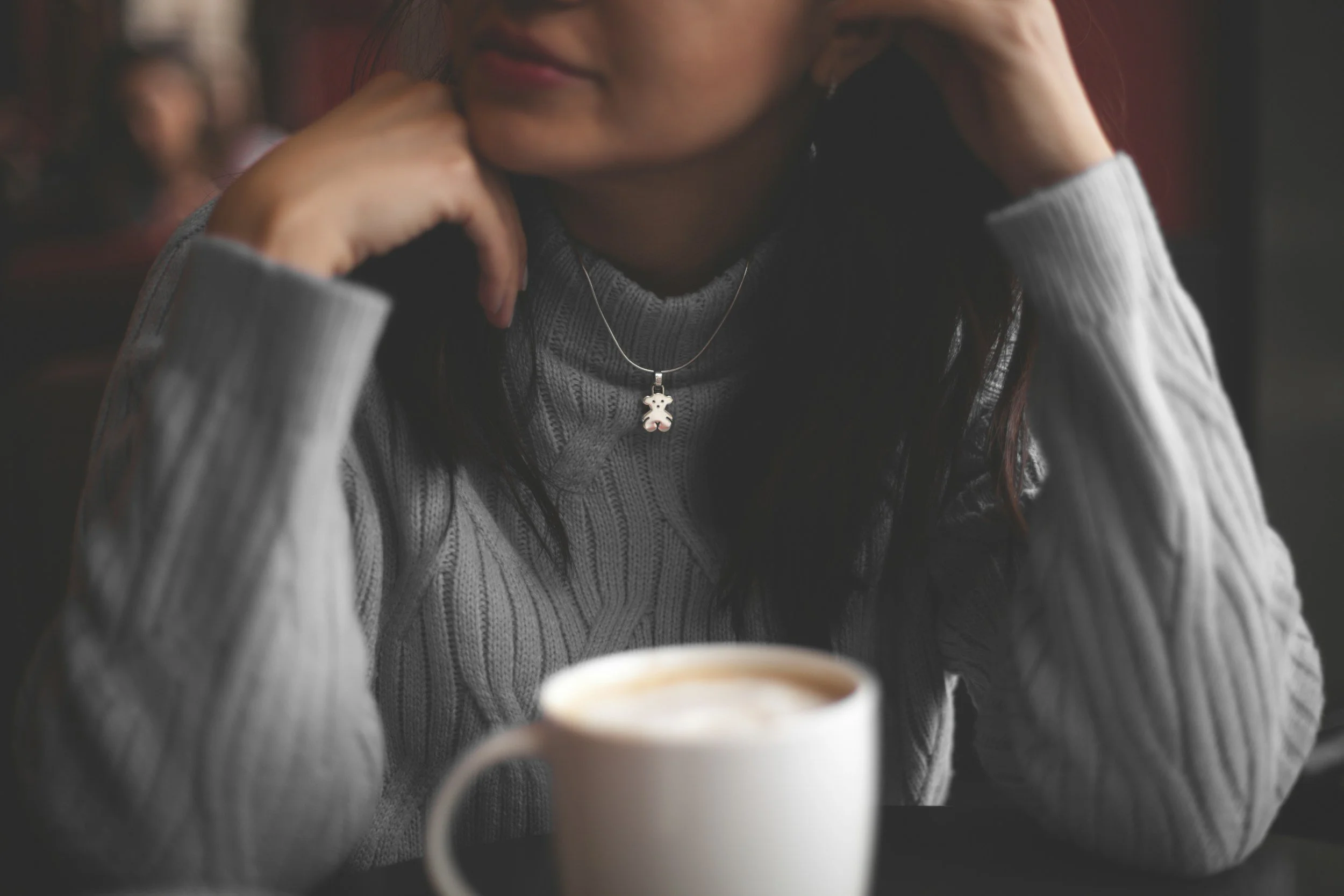 A woman with dark hair wearing a grey cable-knit sweater and a teddy bear pendant necklace, sitting at a table with a white mug of coffee in front of her, resting her chin on her hand, in a cozy cafe setting.