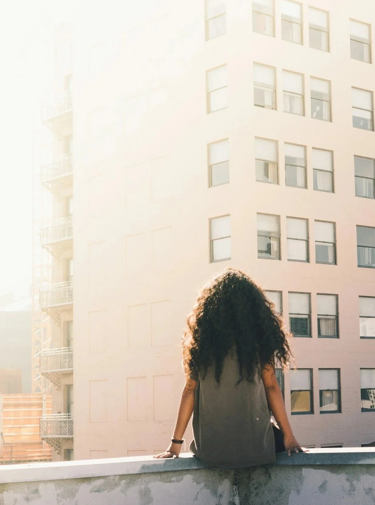 Person with curly hair sitting on a ledge on a rooftop with multi-story apartment buildings in the background, bathed in sunlight.