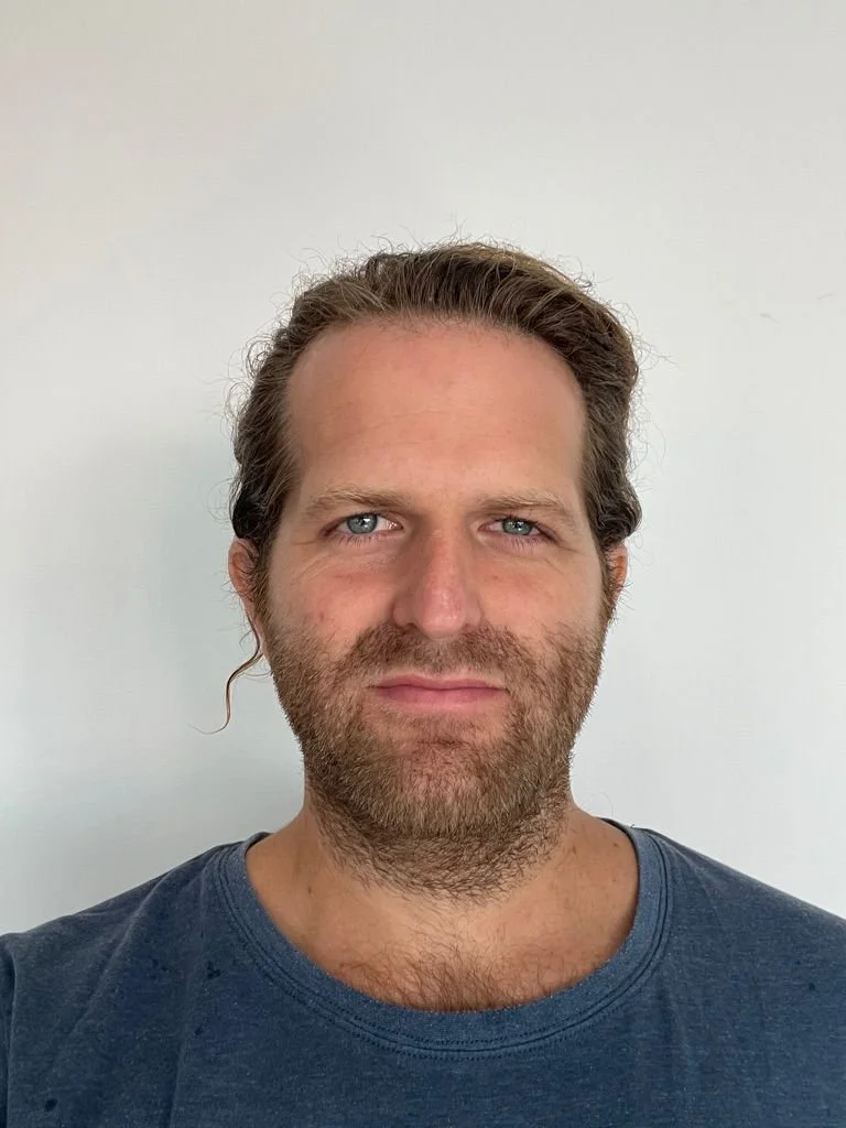 A man with light skin, blue eyes, brown hair, and a beard, wearing a gray T-shirt, standing against a white wall.