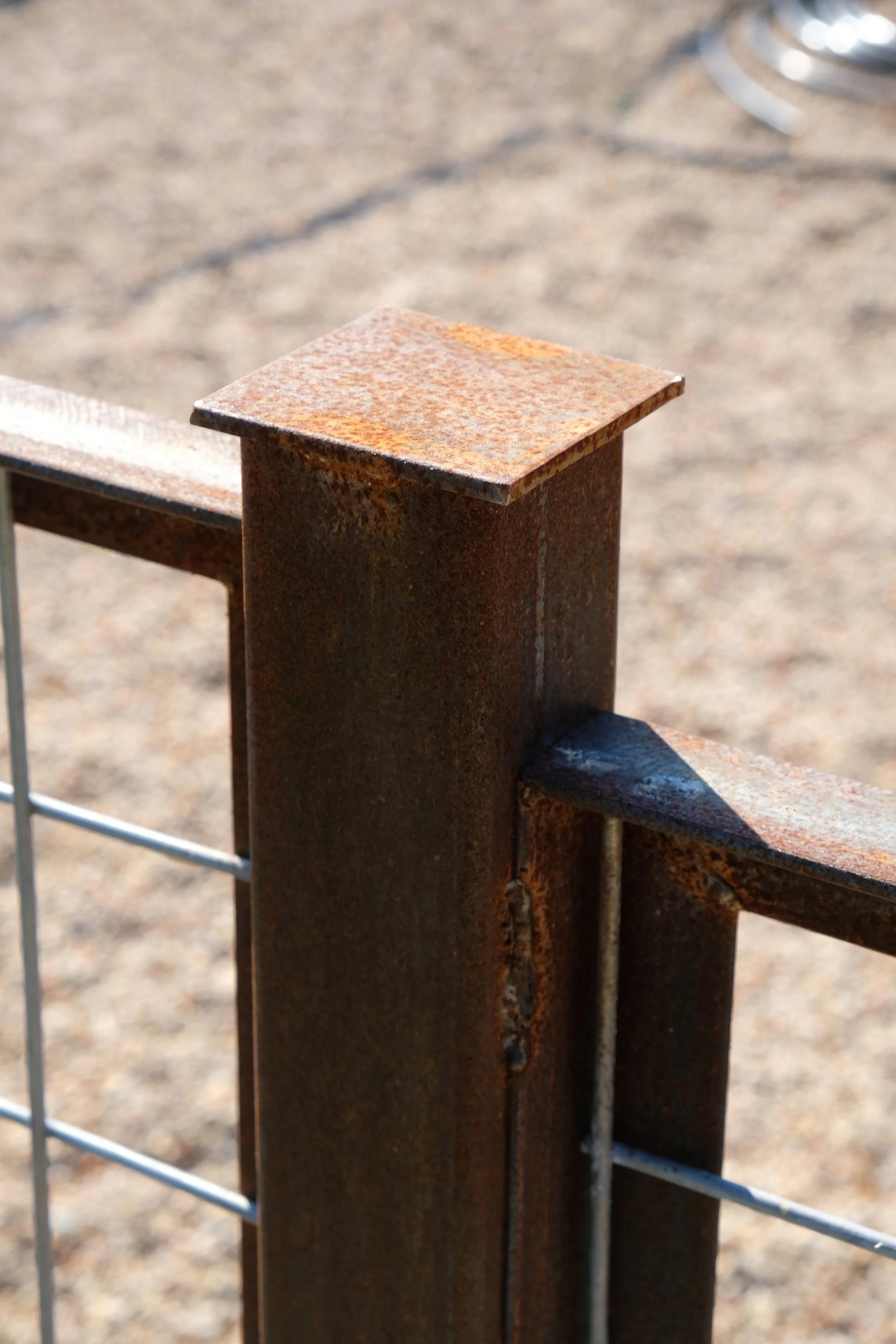 Close-up of a rusted metal fence post with a horizontal bar and wire fencing, on a gravel ground.