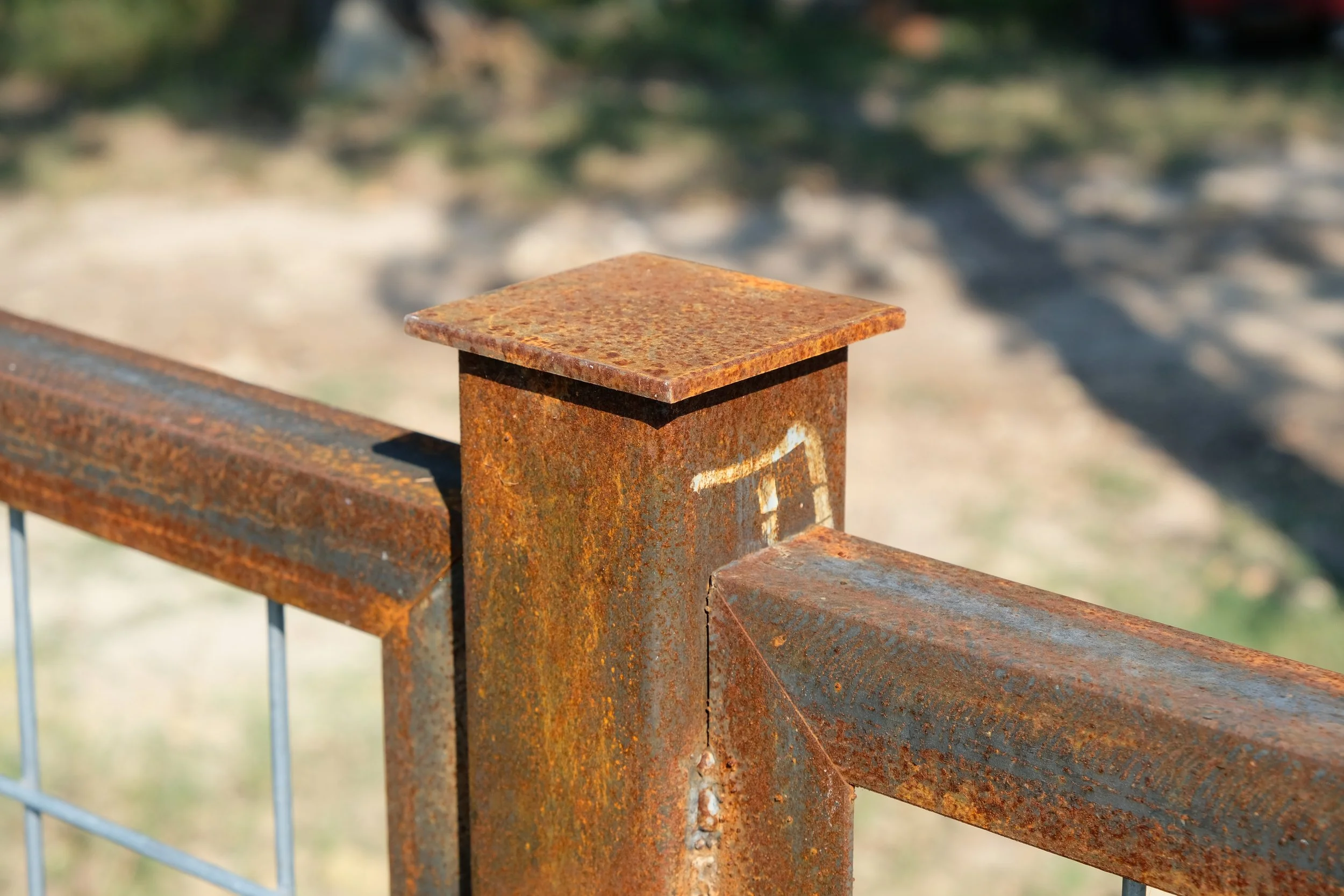 Close-up of a weathered, rusted metal fence post and rail, with a blurred outdoor background.