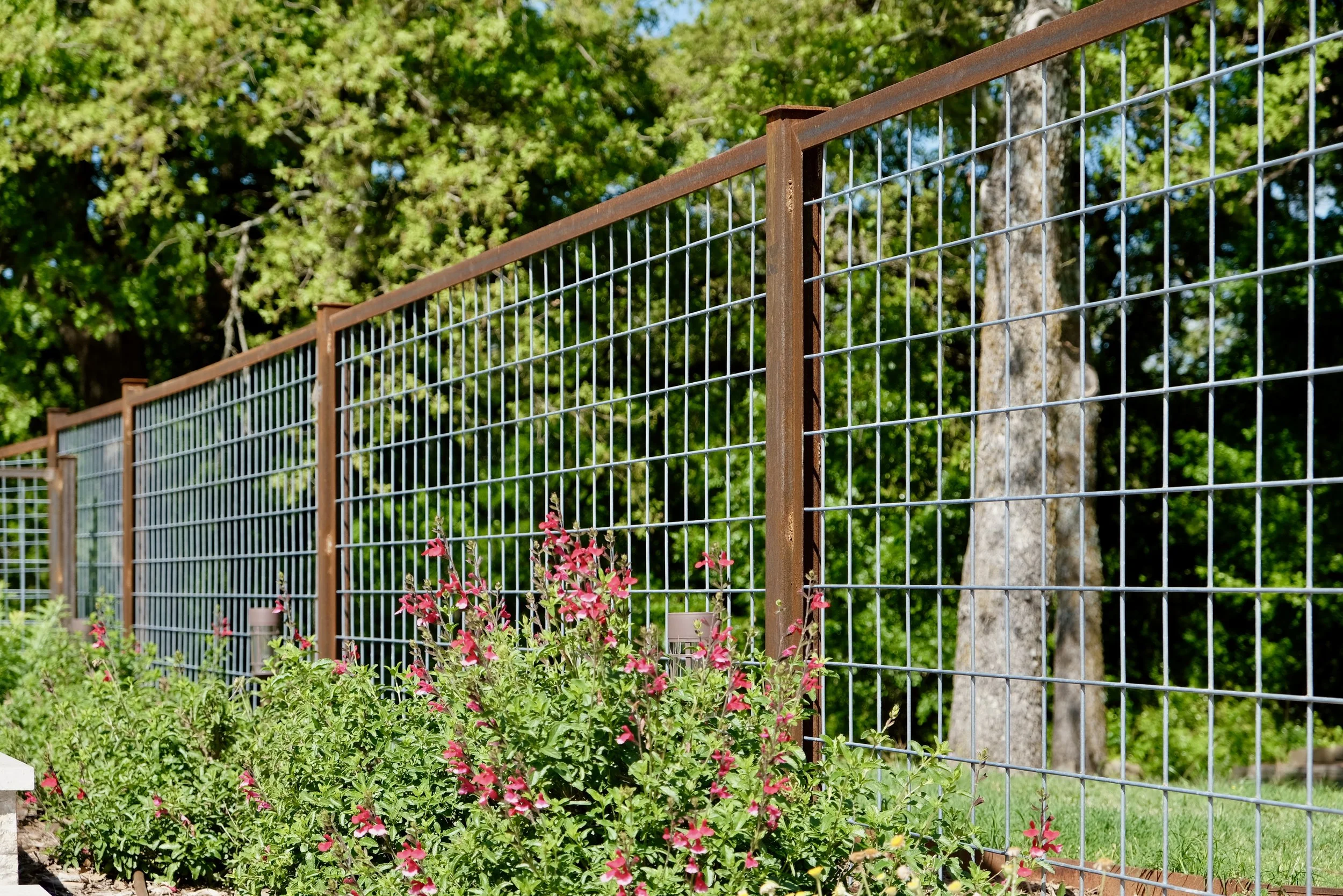 A metal wire fence with rust-colored vertical posts and grid panels runs through a garden with pink flowering plants and green foliage. In the background, there are tall trees with green leaves and a bright blue sky.