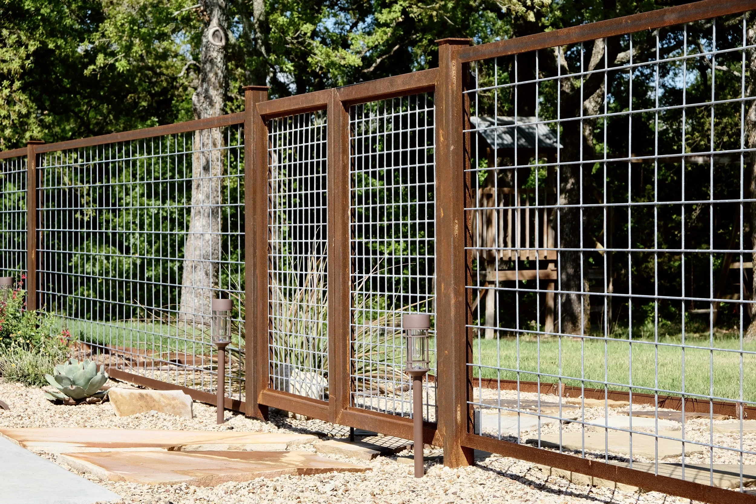 View of a backyard fenced in with rust-colored metal and wire mesh, featuring desert plants and trees, a small light on the ground, and a wooden treehouse in the background.