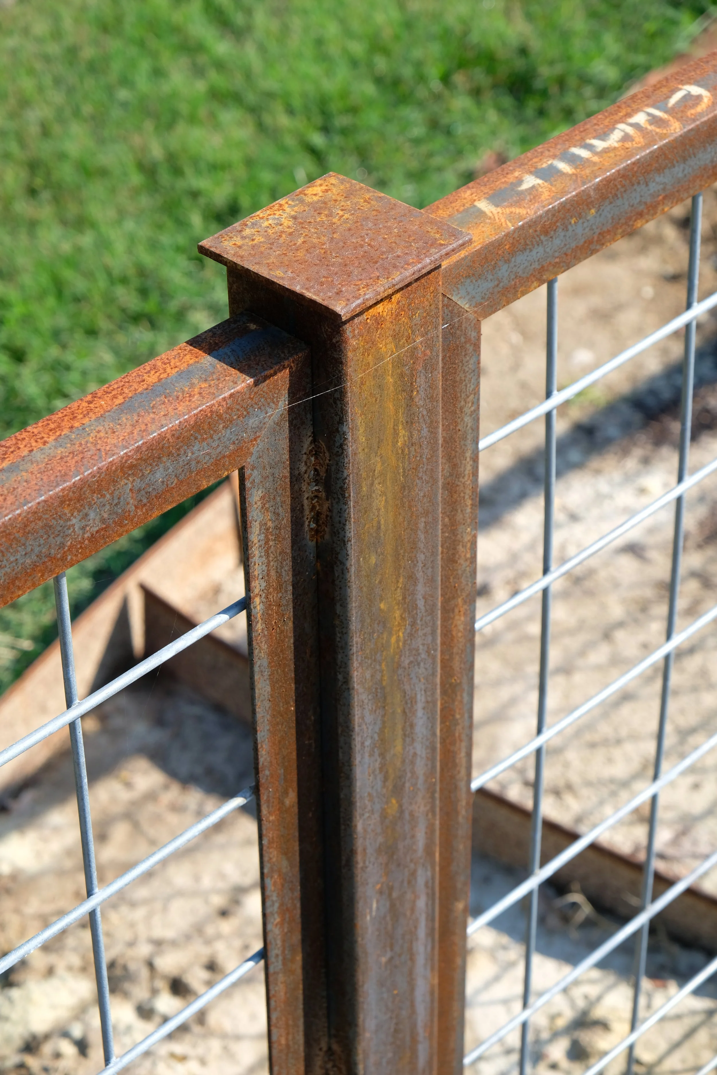 Close-up of a rusted metal fence post with a rusty top cap, attached to a wire grid fence, with a grassy area and dirt ground in the background.