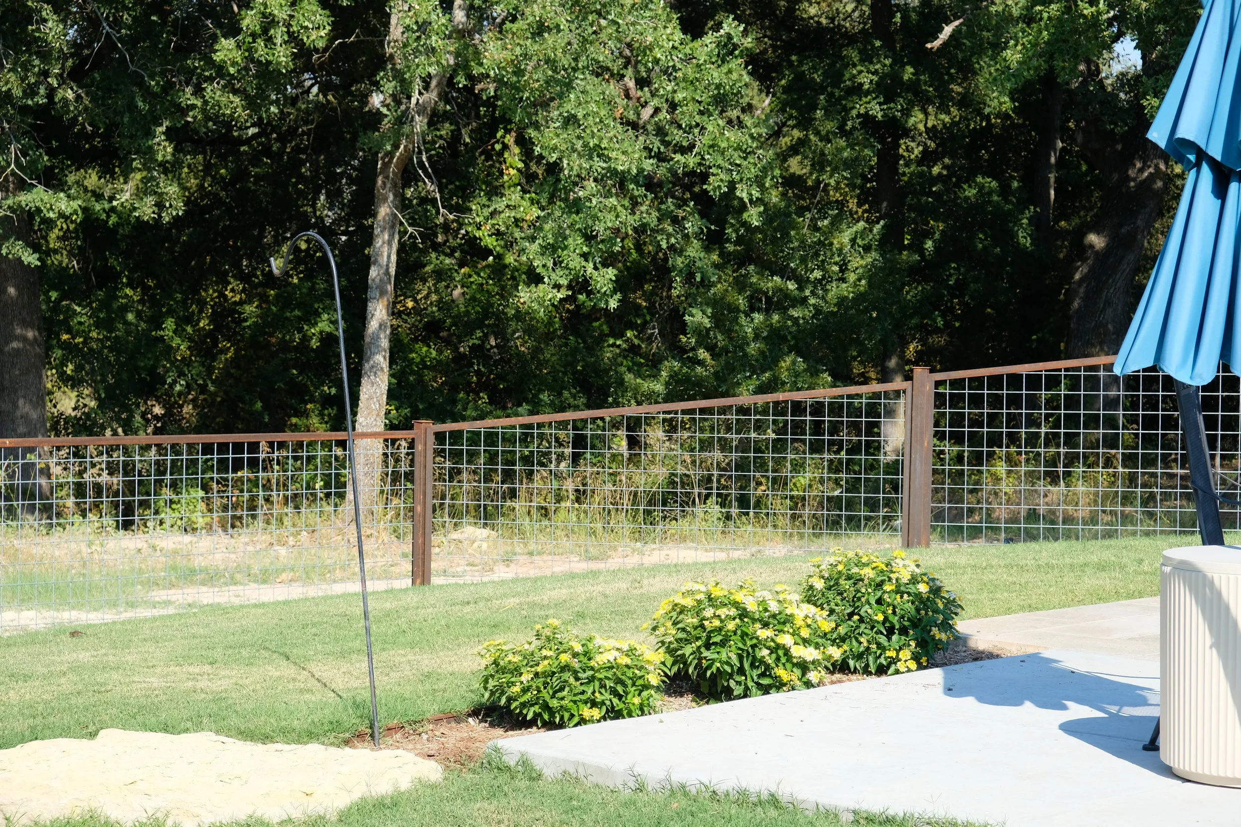 Backyard with a grassy lawn, a wire fence, on which a black outdoor shower is installed, and a concrete patio with a blue patio umbrella, plants, and trees in the background.