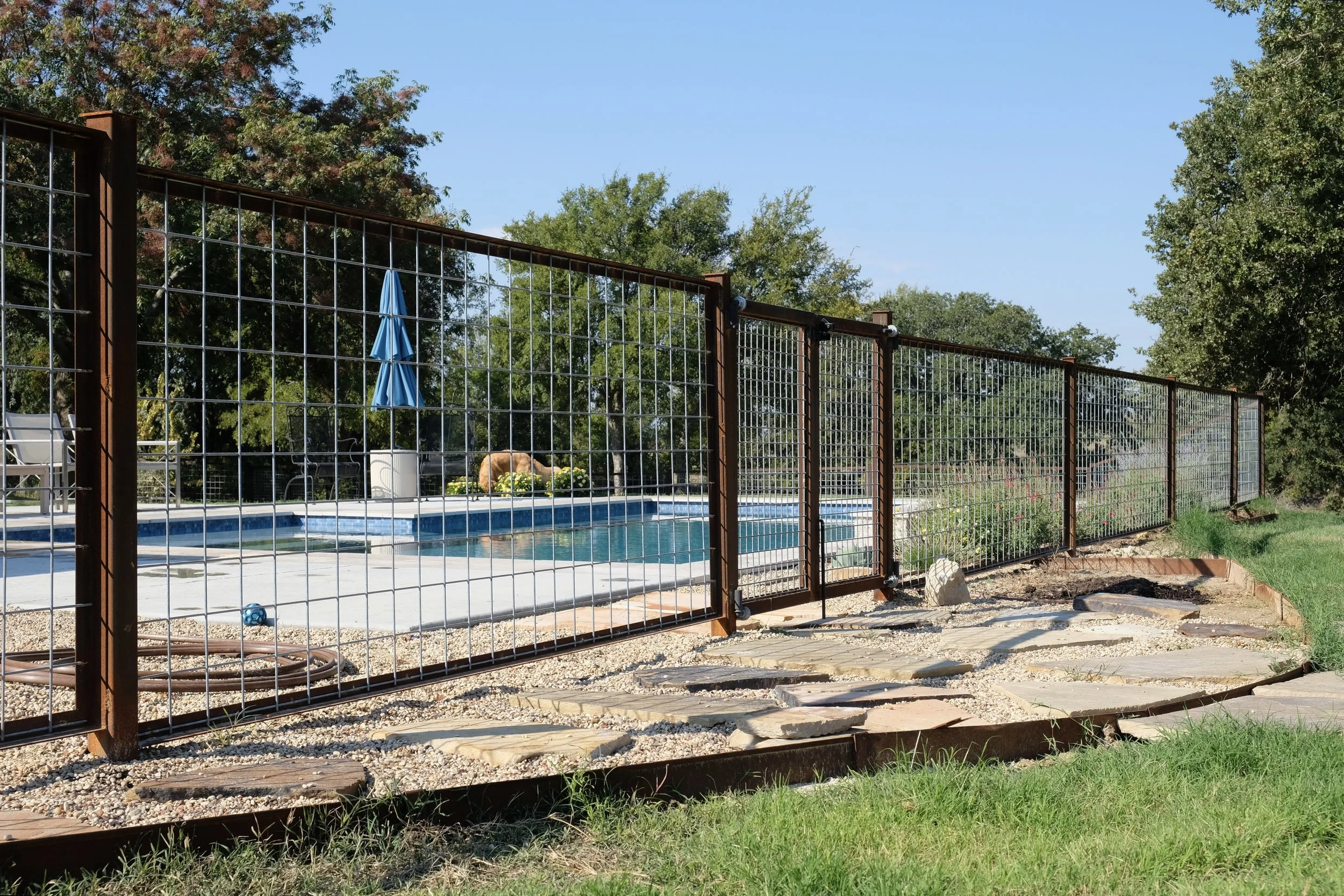 Backyard with a pool enclosed by a metal fence, surrounded by green trees and shrubbery, blue sky overhead.