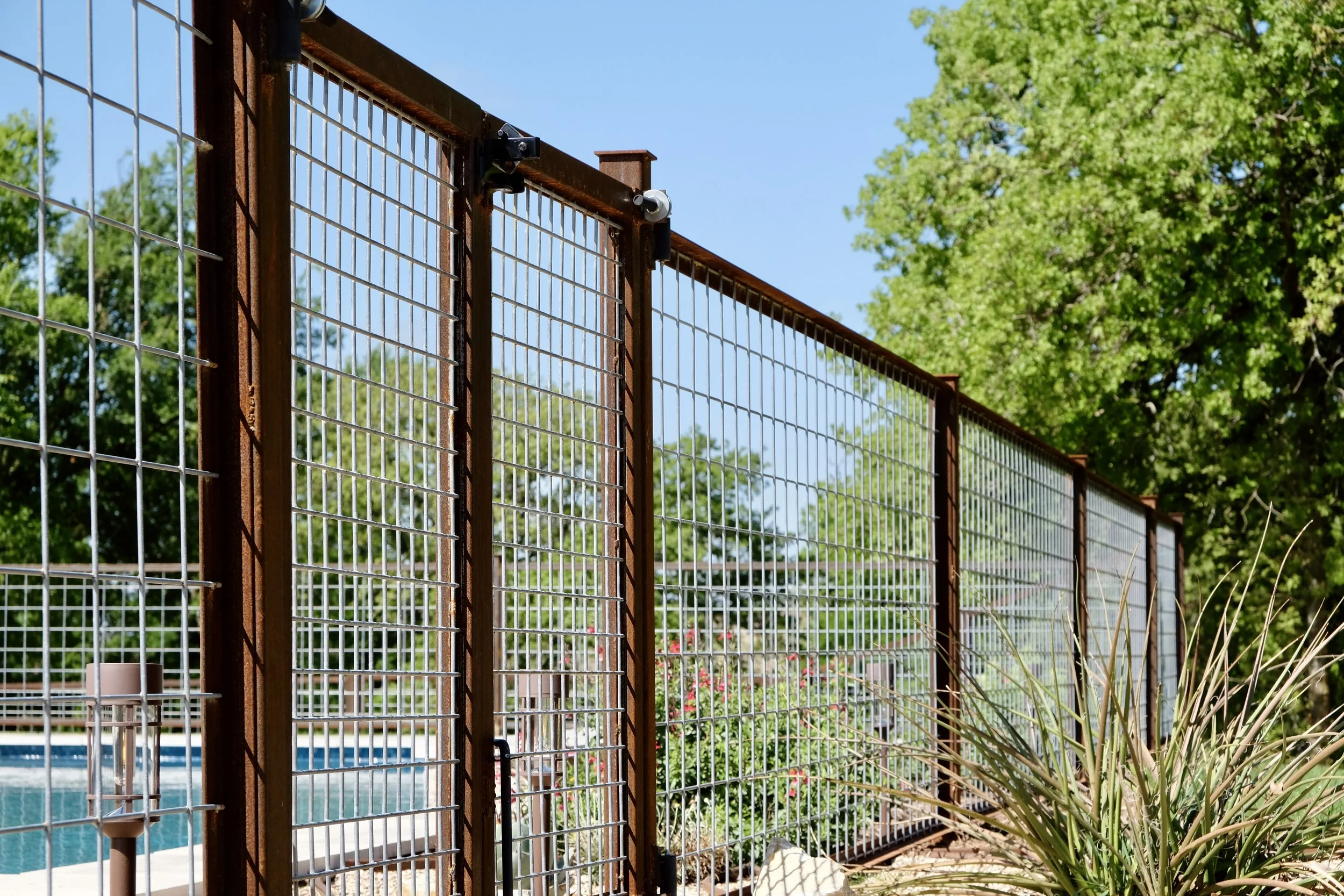 A metal fence with rust finish and wire mesh panels, part of a pool enclosure, on a clear sunny day with green trees and plants in the background.