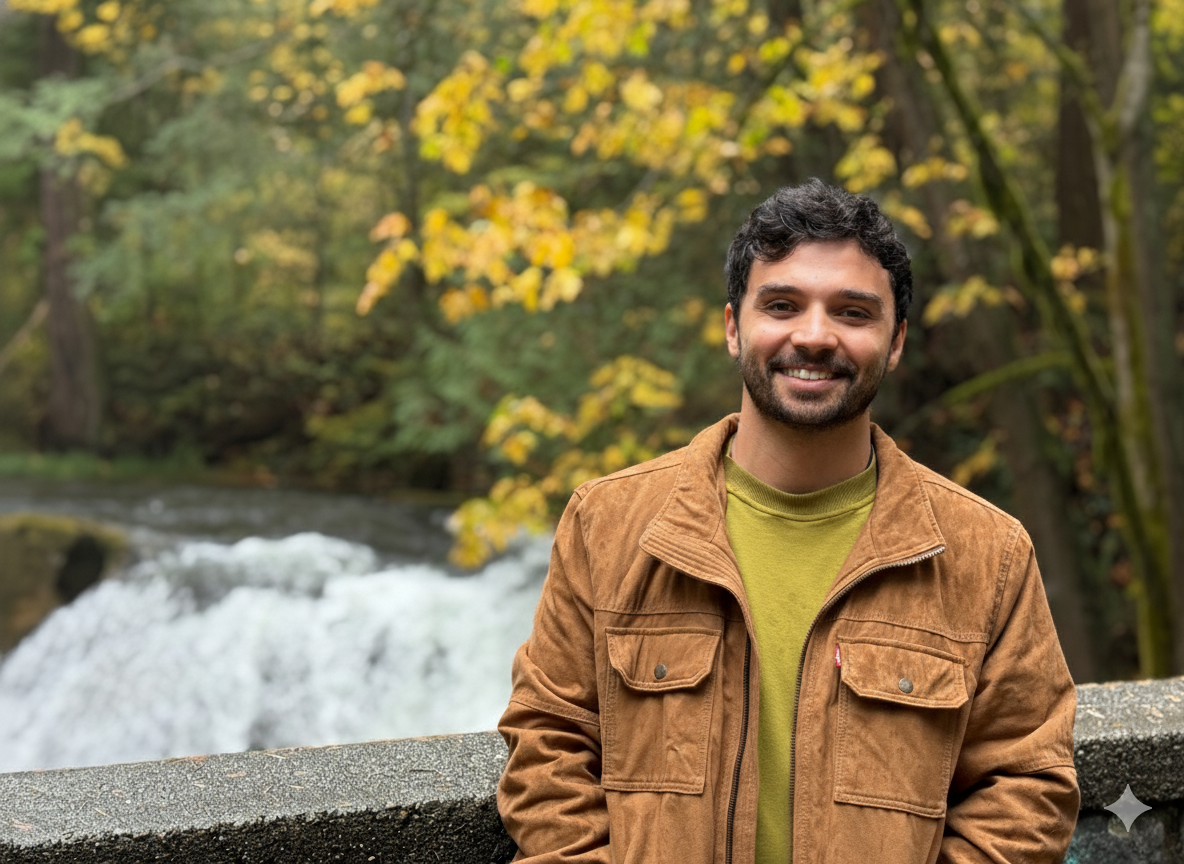 A smiling man with dark hair and a beard stands outdoors in front of a waterfall and trees with yellow and green leaves. He is wearing a brown jacket over a green shirt.