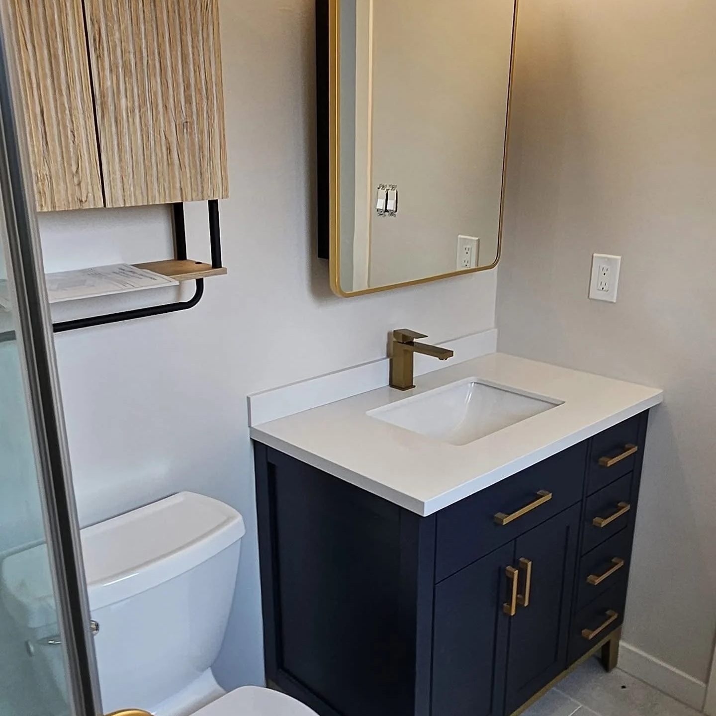 Modern bathroom vanity with dark blue cabinet, brass handles, white countertop, rectangular sink, brass faucet, wall-mounted mirror, wooden shelf, and toilet in a small space.