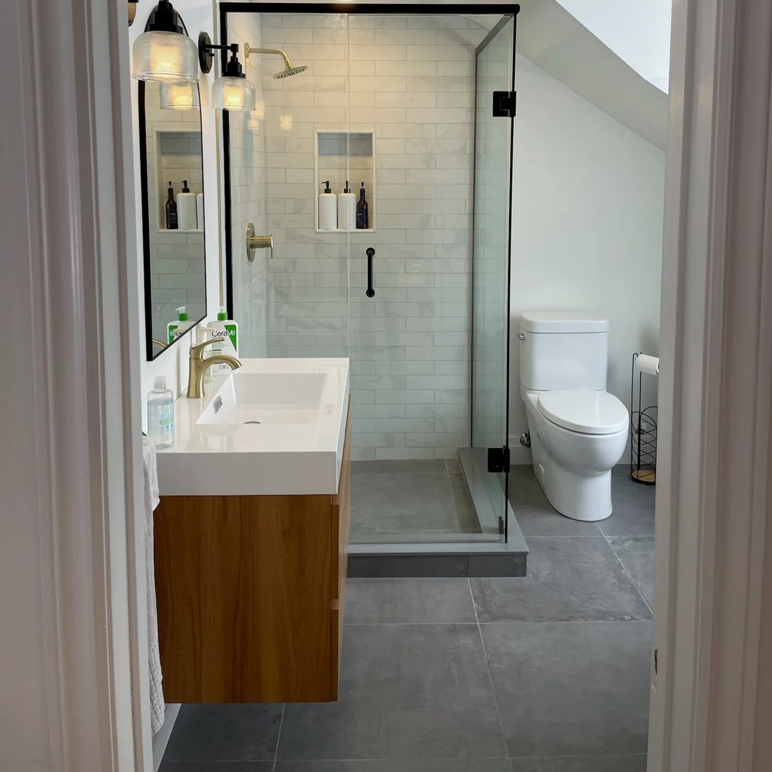 A modern bathroom with a glass shower enclosure, white subway tile walls inside the shower, a white countertop with a wooden cabinet, a gold faucet, a mirror, a toilet, and a small black wire trash can.