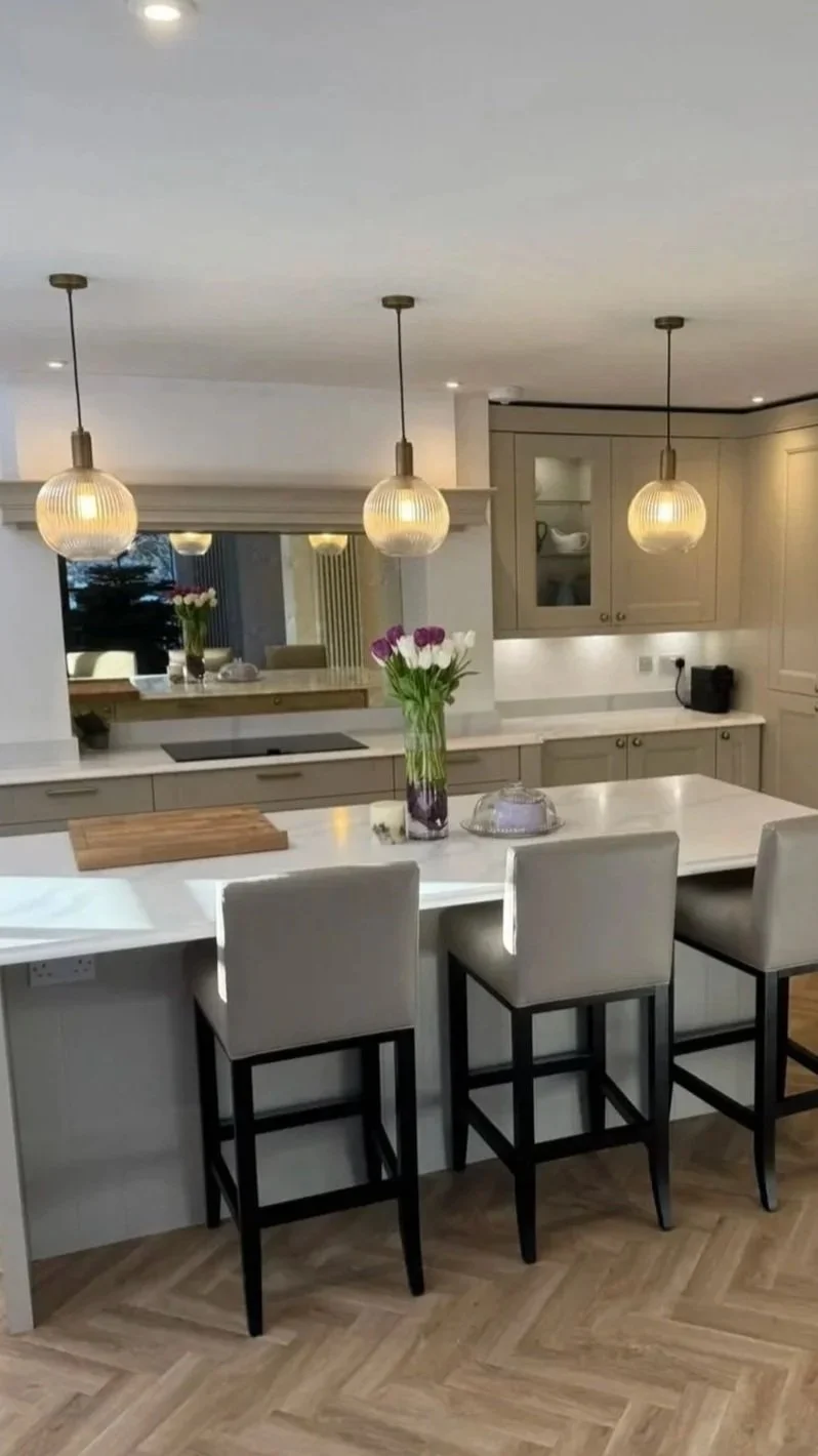 Modern kitchen with a white island, beige cabinets, three pendant lights, and a vase with purple and white flowers on the island.