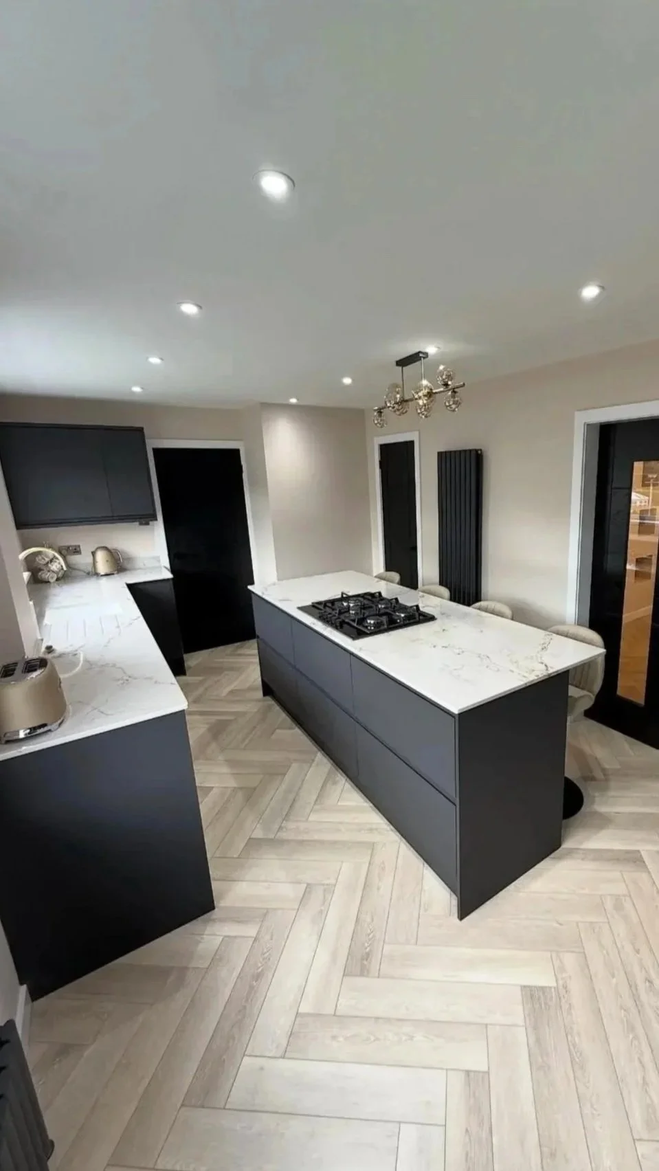 Modern kitchen with black cabinets, white marble countertops, a kitchen island with a gas stove, and light wood herringbone flooring.