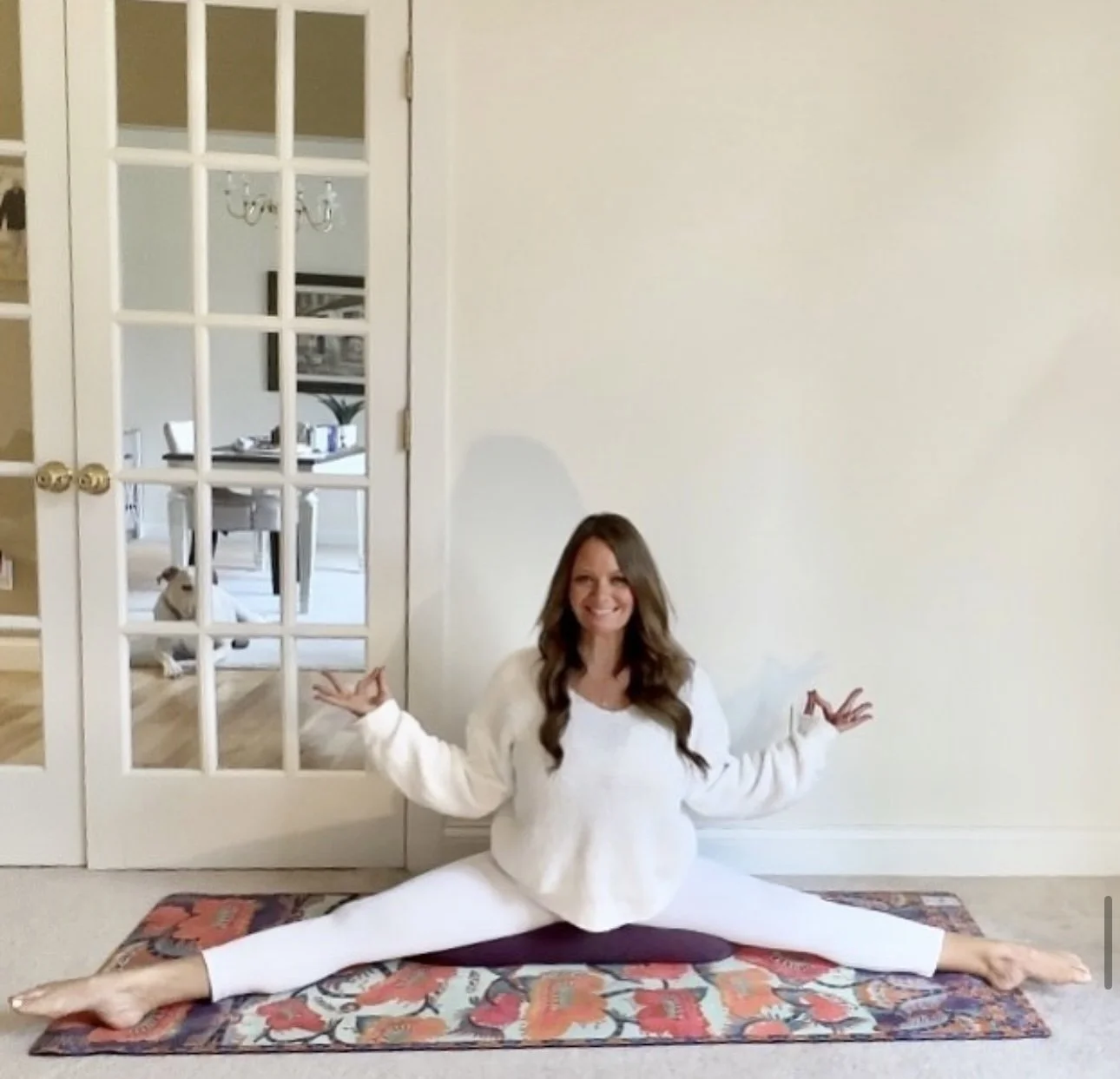 Woman in white sweater and white leggings doing a split yoga pose on a colorful rug in front of a plain wall, with a glass-paneled door and a dog sitting inside the room behind her.