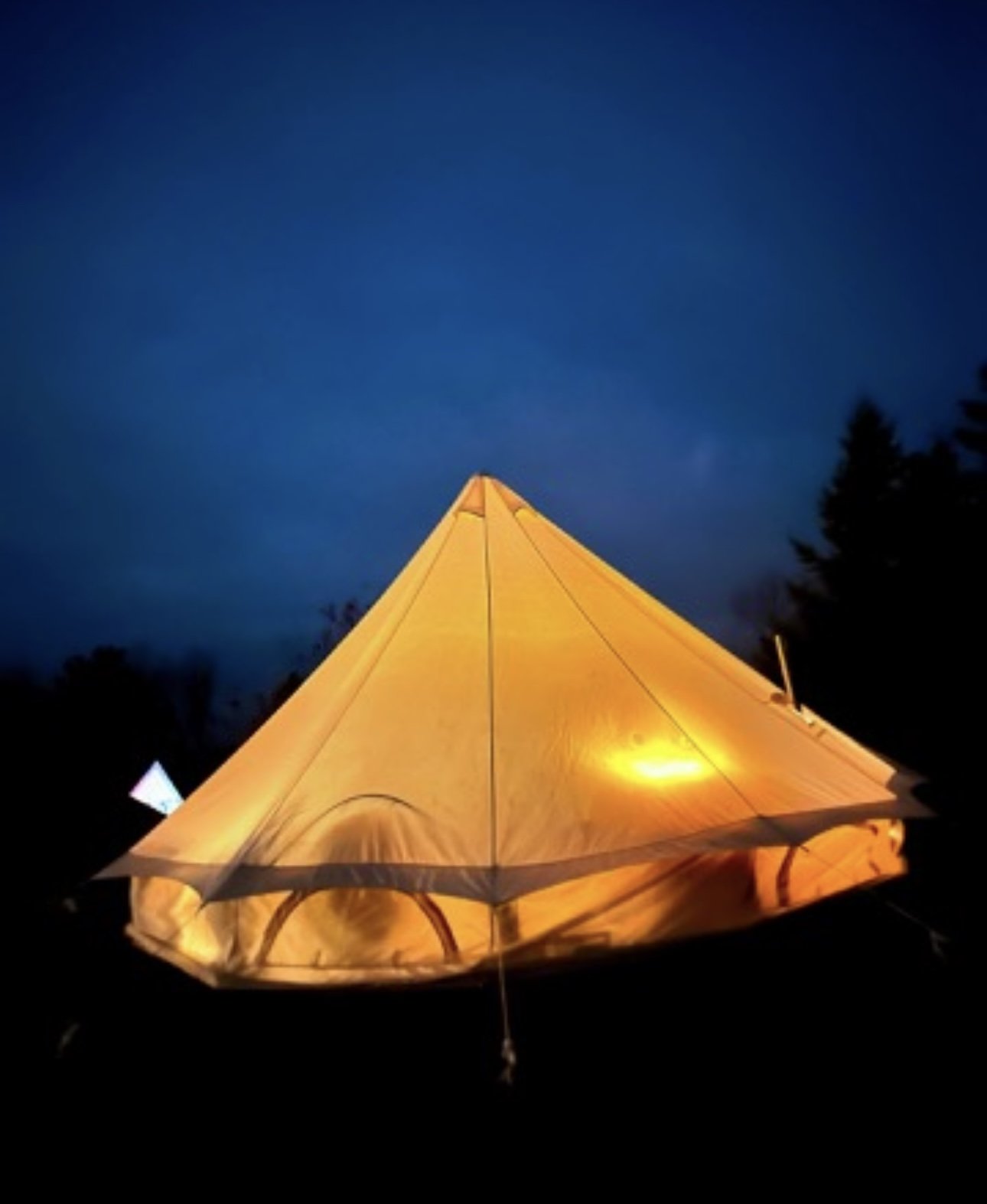 A yellow tent illuminated from within at night, set against a dark blue sky with silhouettes of trees in the background.