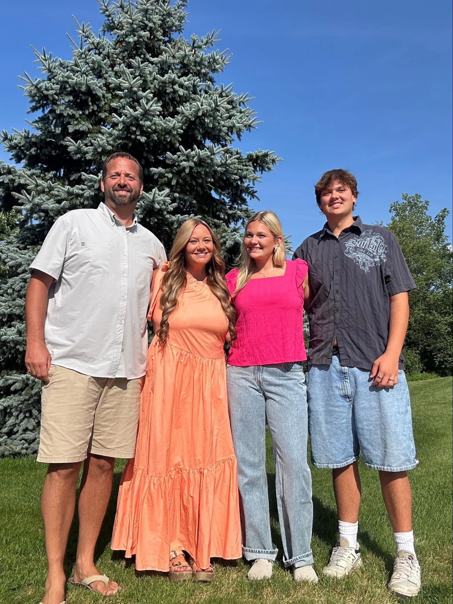 Four people standing outdoors in front of a large evergreen tree, smiling, under a clear blue sky.