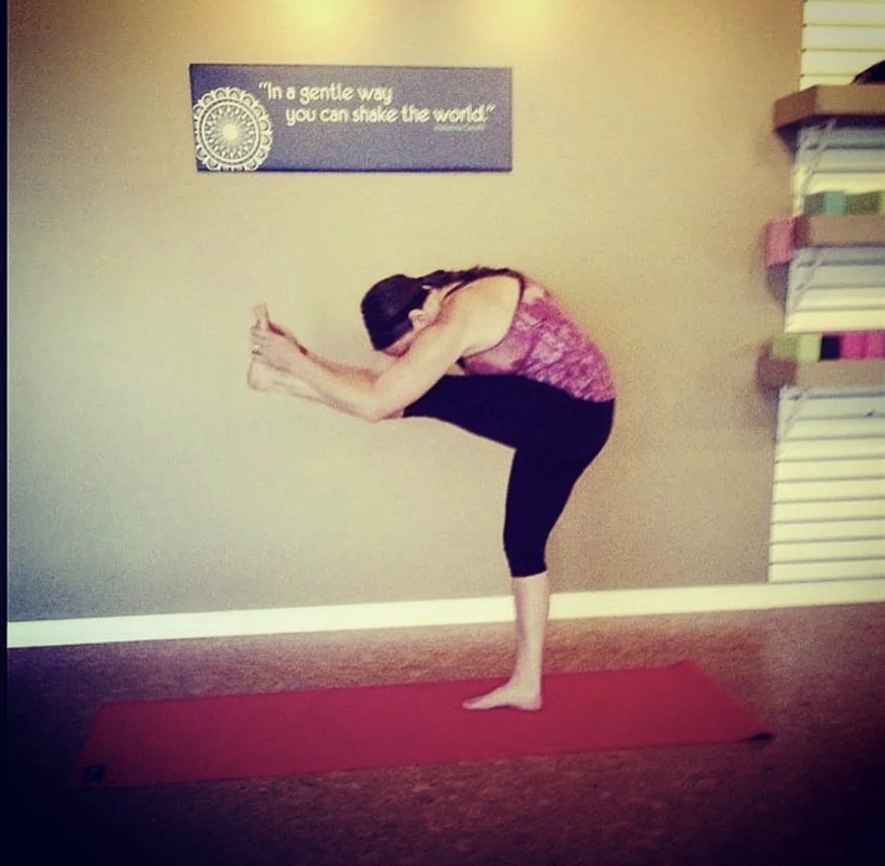 Woman practicing yoga in a yoga pose on a red mat in a room with beige walls, a bookshelf, and a framed quote that says 'In a gentle way you can shake the world.'.