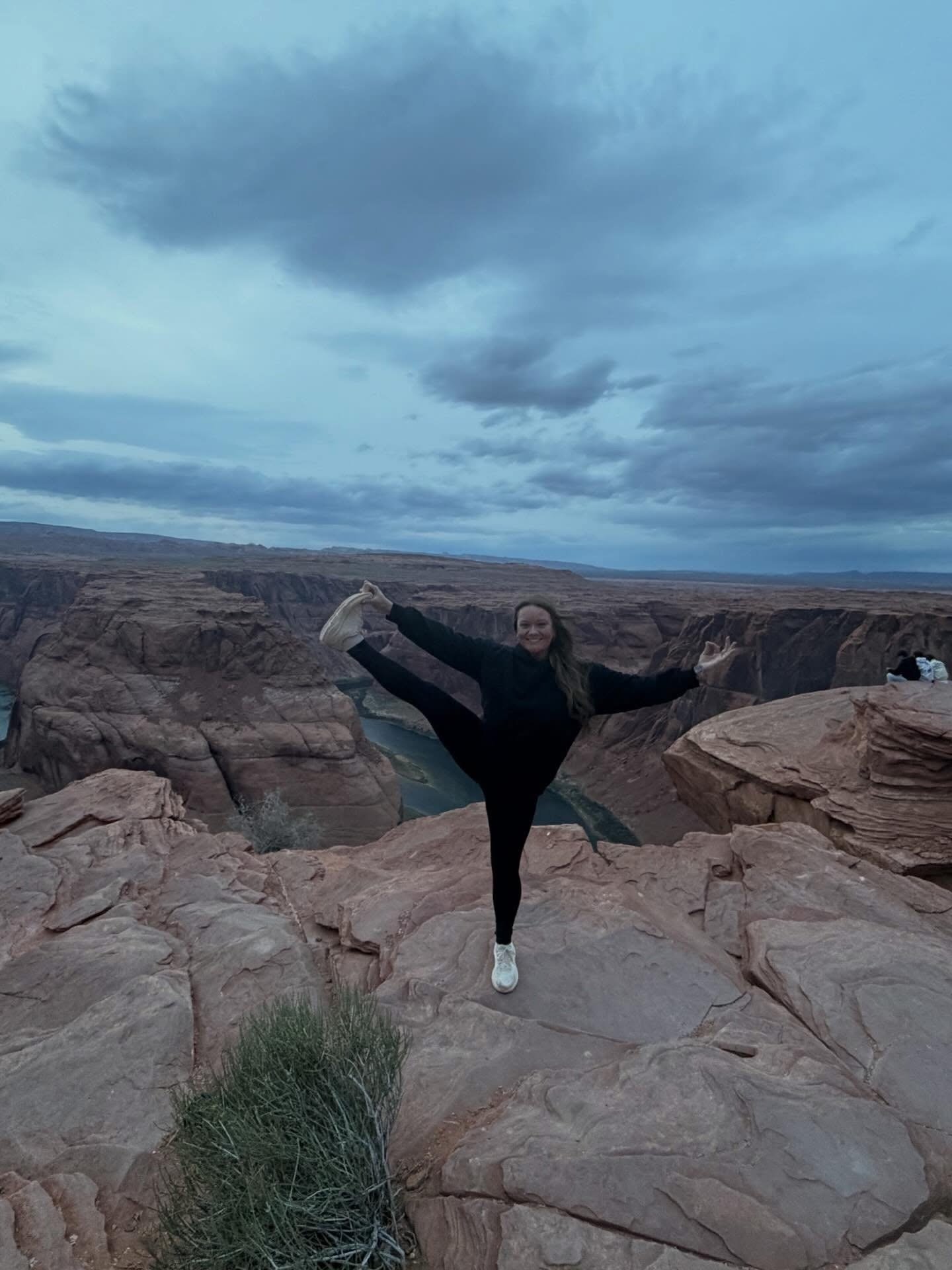 A woman in black athletic clothing and white sneakers is balancing on one leg in a desert canyon landscape, with her other leg extended upward and her arms outstretched.