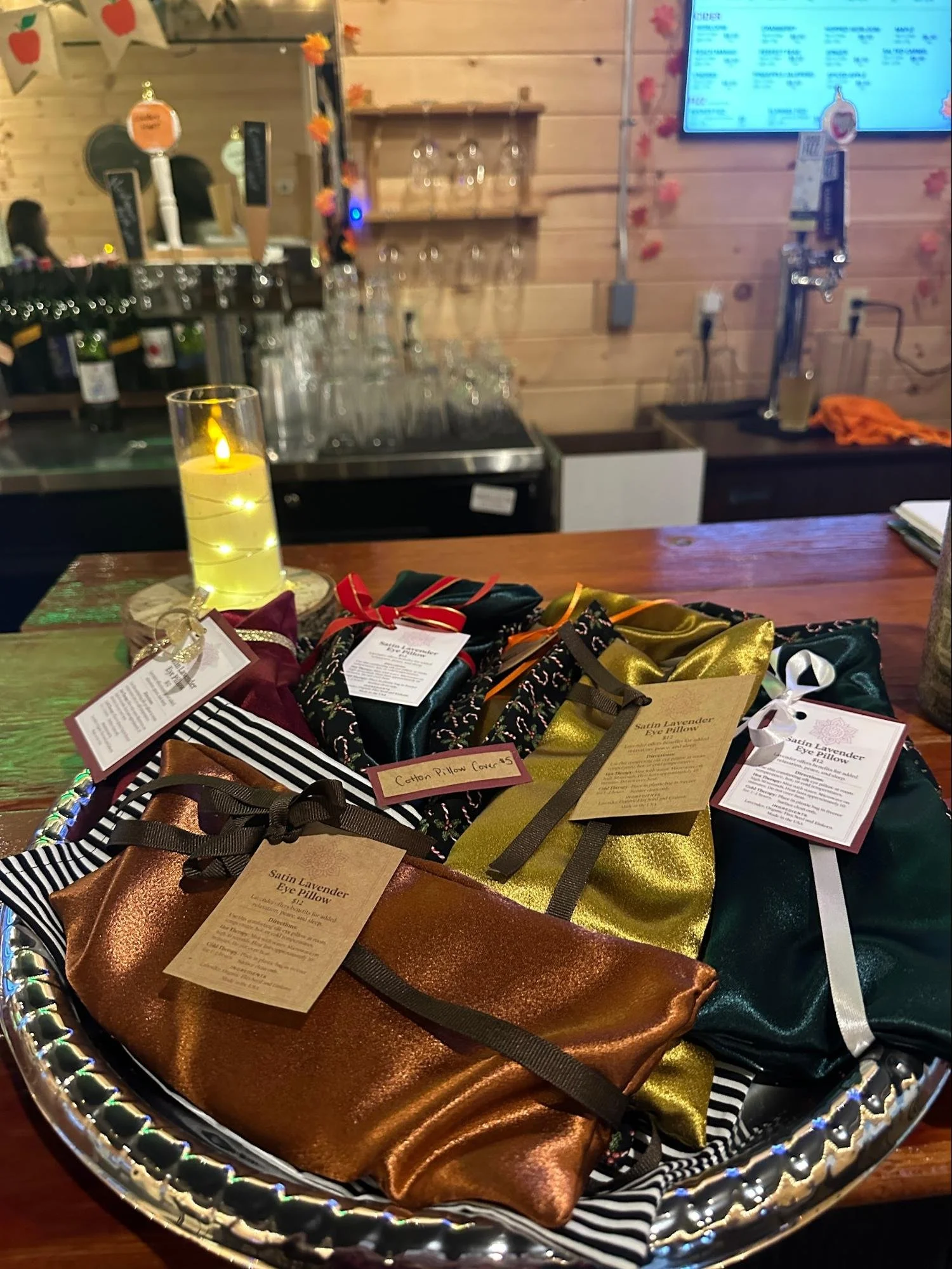 A silver tray holding several satin lavender eye pillows wrapped with ribbons and price tags, with a cozy bar or restaurant background featuring glasses, a digital menu screen, and wooden wall decor.