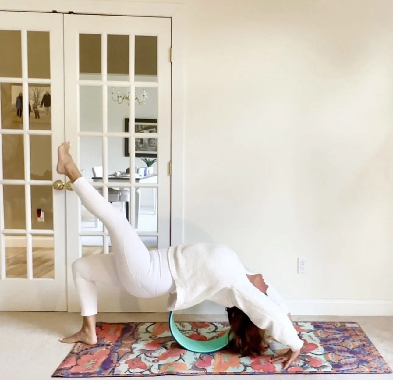 Person in a white robe performing an upside-down yoga pose on a colorful rug in a living room, with the head and hands on the floor and one leg raised straight up, in front of a glass door.