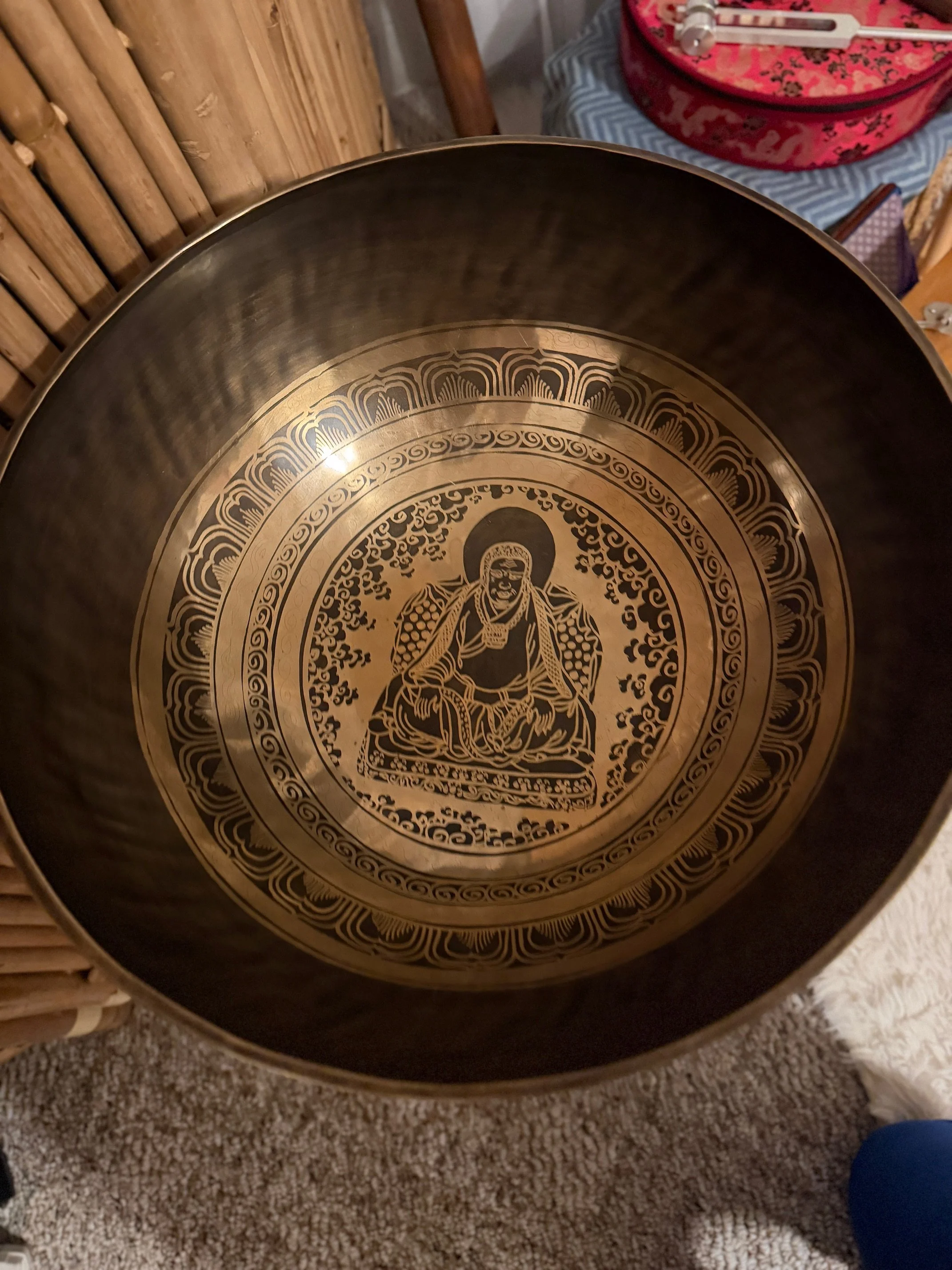 Close-up of a metallic singing bowl with intricate black and gold design on the inside, depicting a seated figure surrounded by decorative patterns.